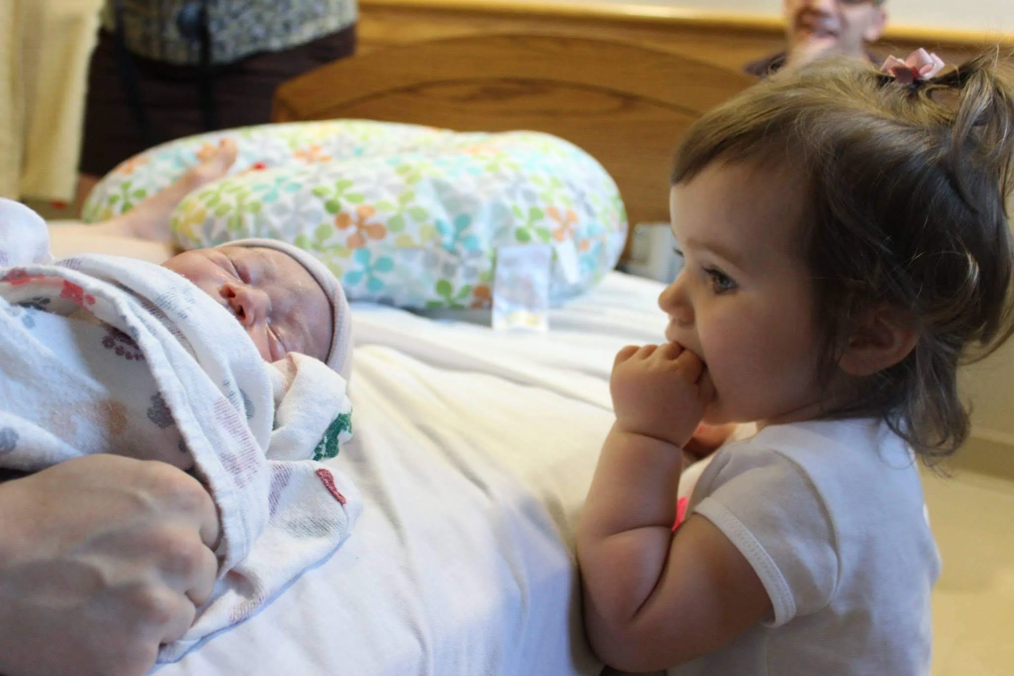 A young girl watching a sleeping newborn baby in a hospital bed as a man, possibly the father, stands in the background.