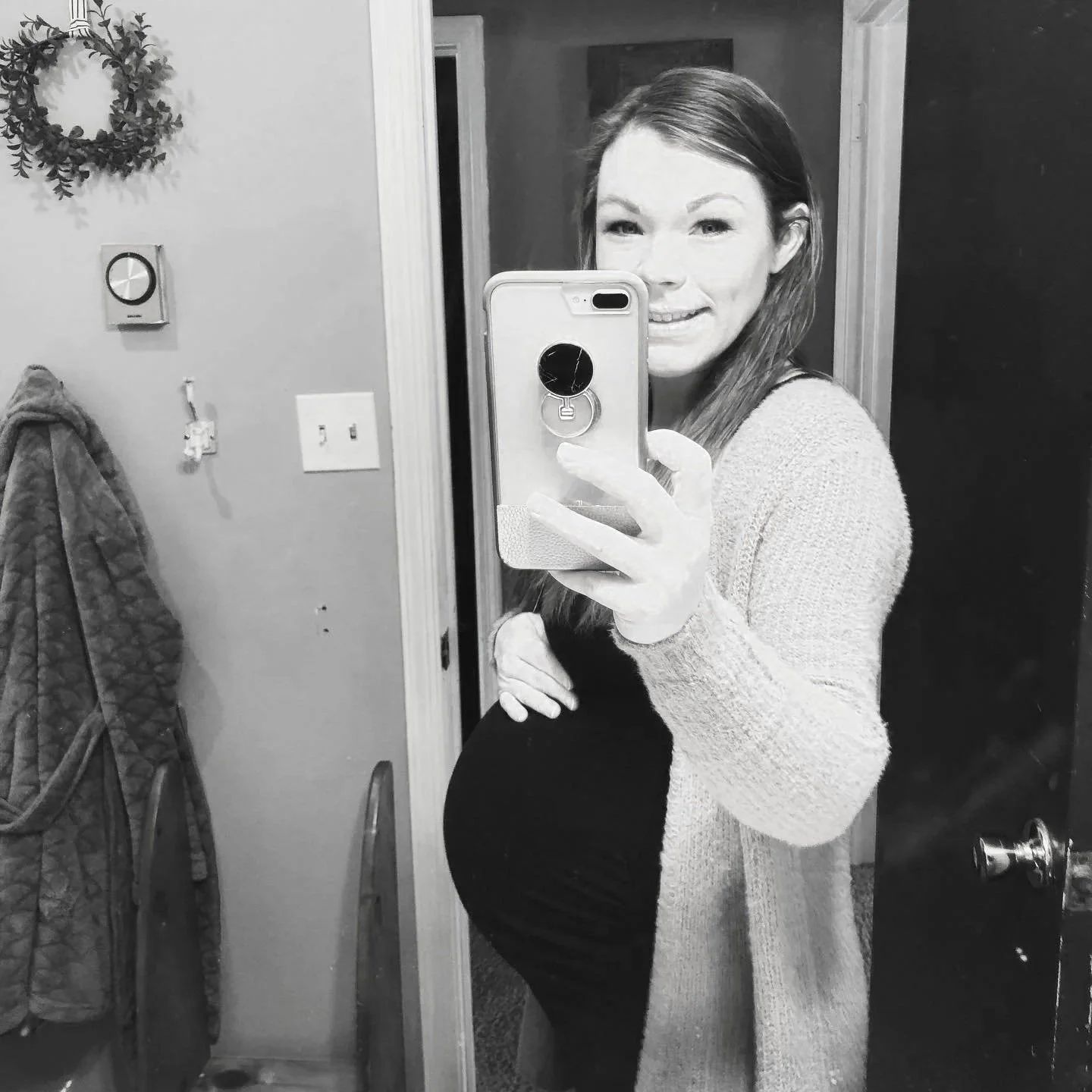 A woman with long hair taking a mirror selfie in a bathroom, showing a pregnant belly, dressed in a black top and cardigan.