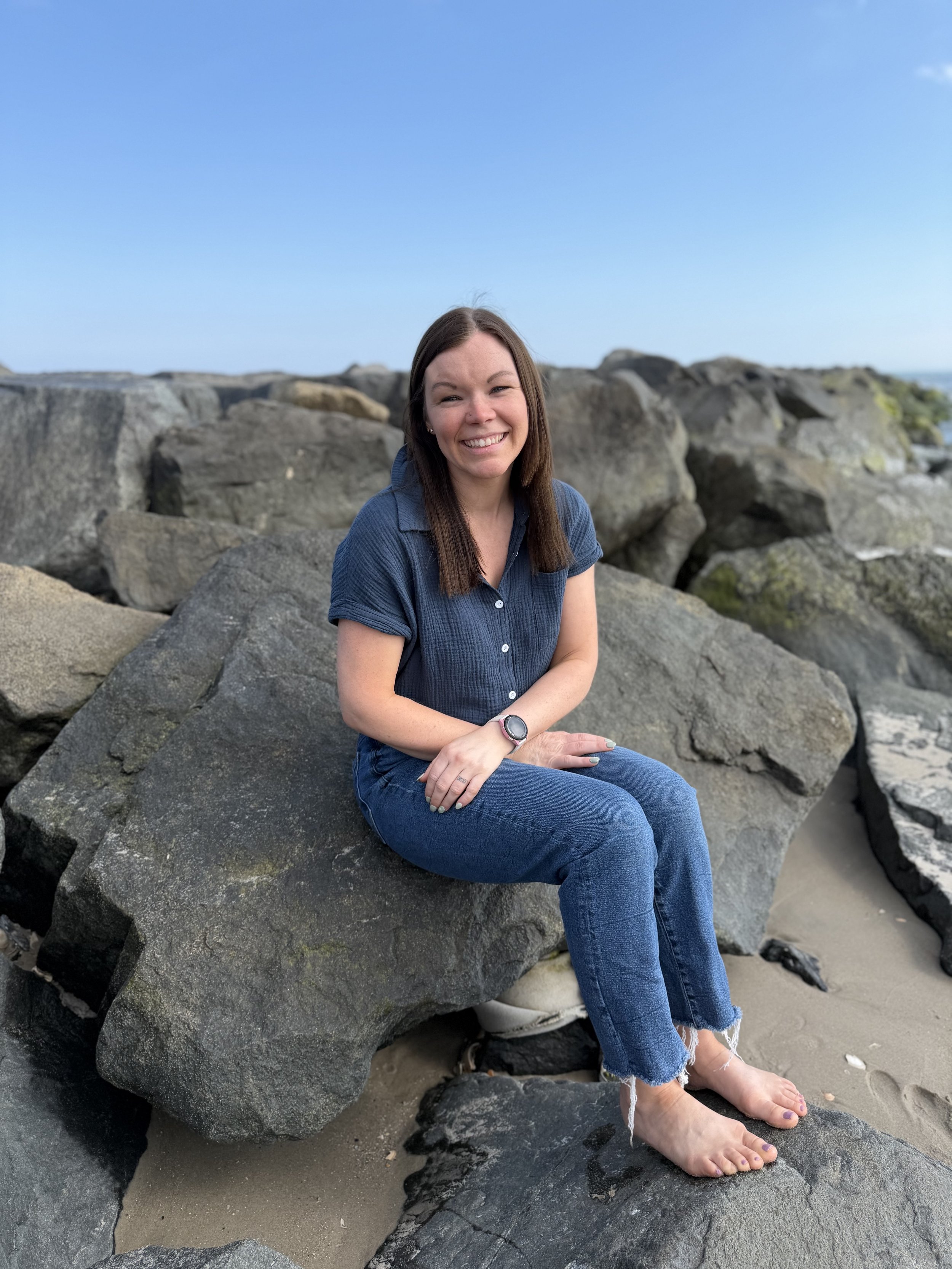 A woman sitting barefoot on a large rock at the beach with rocks in the background and a clear blue sky.