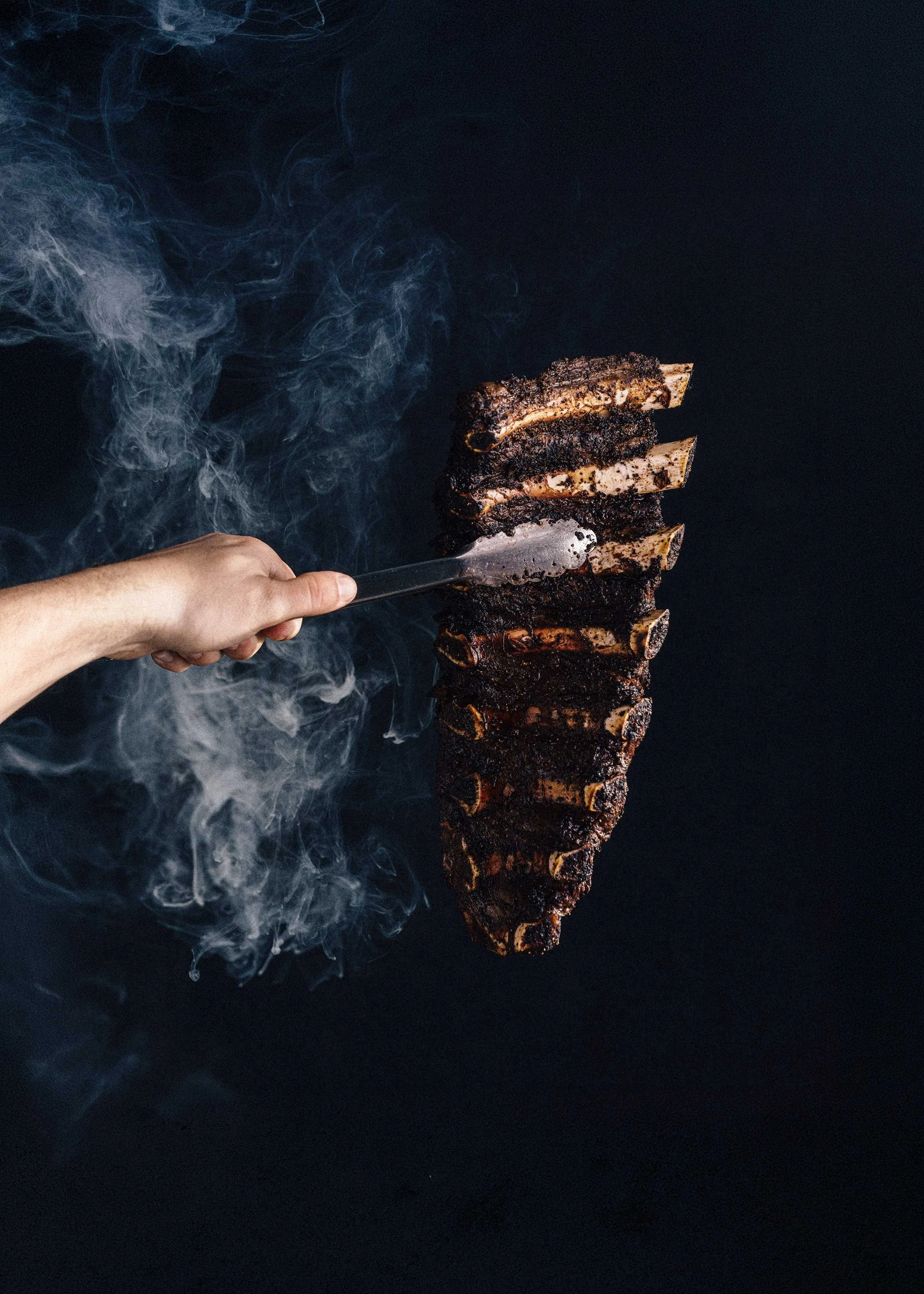 A person holding tongs with meat ribs. The ribs are blackened and smoky, hanging vertically against a dark background with smoke around.