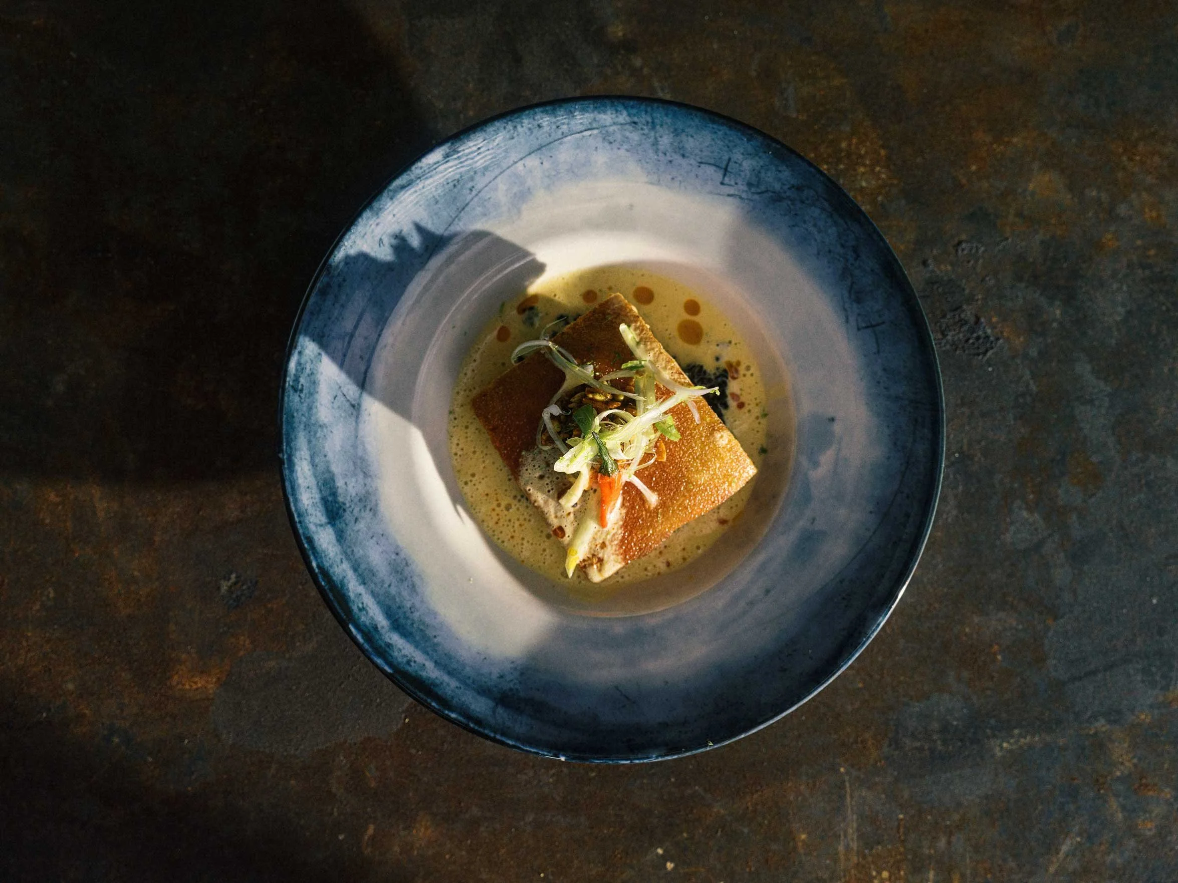 A plated dish with a piece of fried fish topped with microgreens, served in a bowl with a creamy yellow sauce and garnished with red pepper flakes.