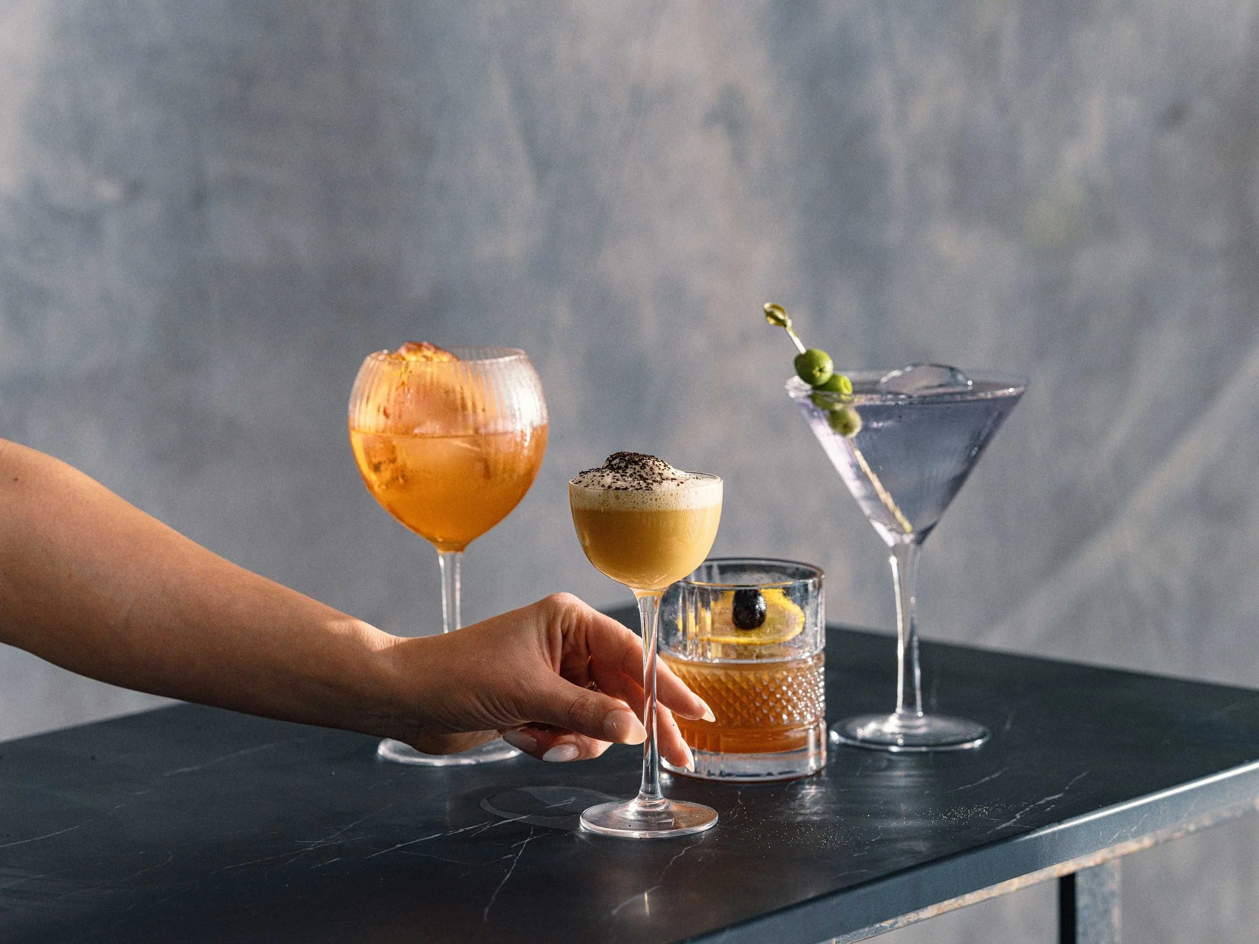 hand holding cocktail glasses including an old fashioned, martini, and citrus drink on a bar