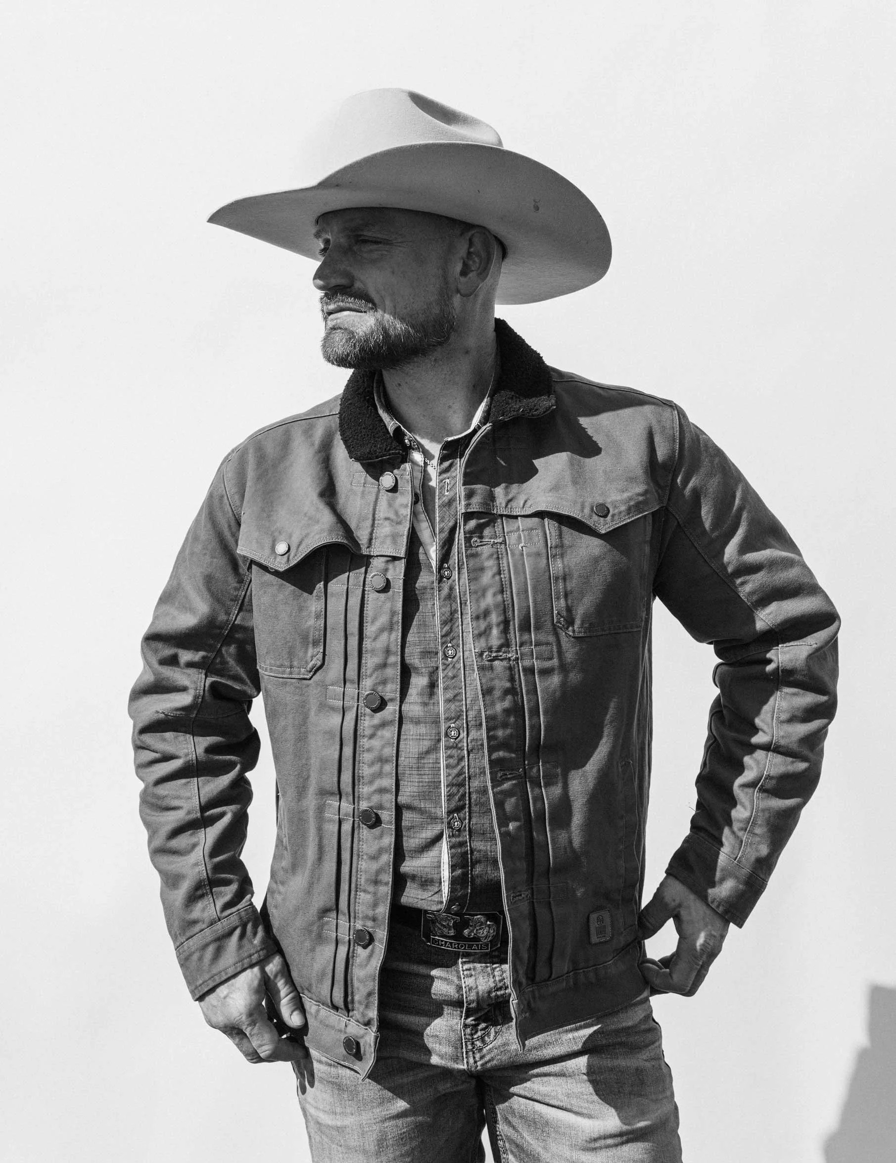Black and white photo of a man wearing a cowboy hat, denim jacket, and jeans, standing against a plain background.