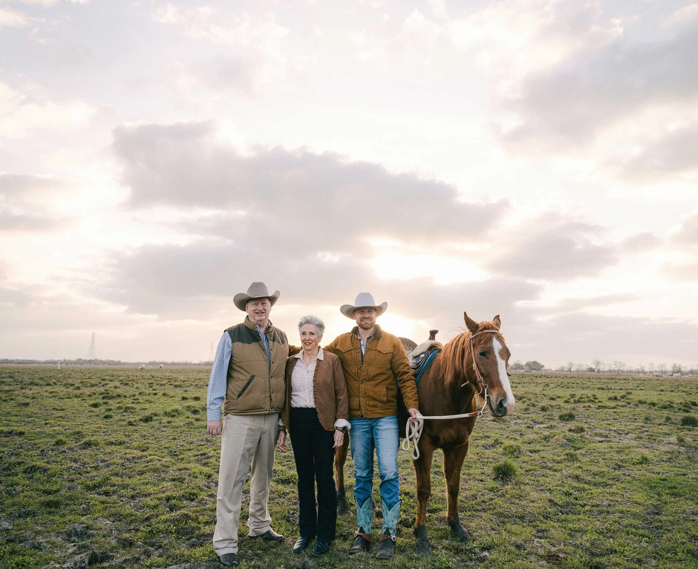 Three people standing in an open field with a horse, all wearing cowboy hats, with a cloudy sky and sunset in the background.