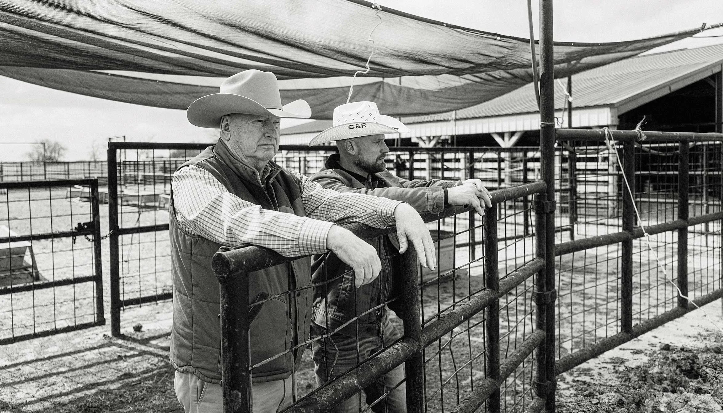 two ranchers leaning on a fence in a cattle yard black and white