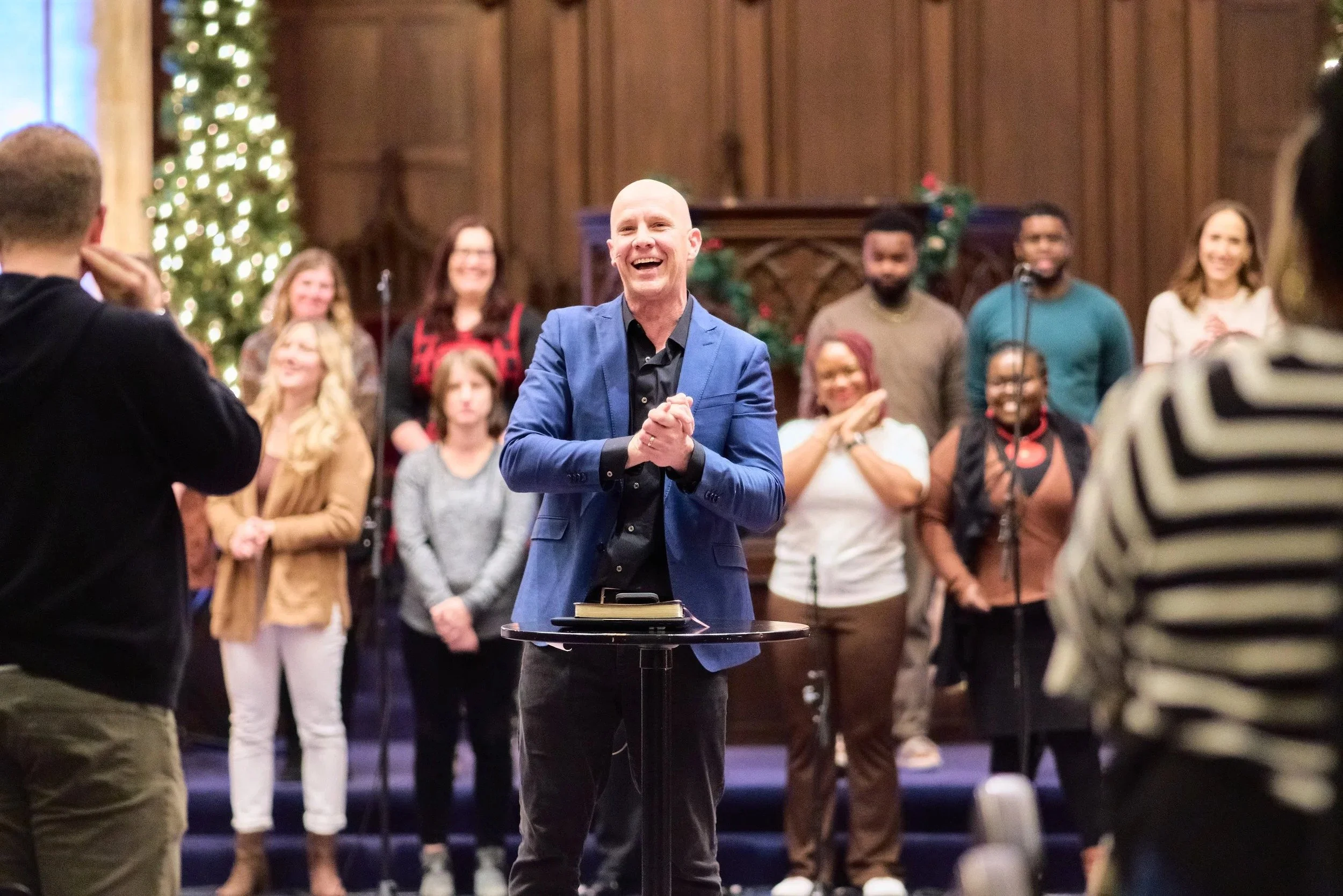 A man in a blue blazer and black shirt smiling on stage, surrounded by a group of people, some smiling and others clapping, with a Christmas tree and church interior in the background during a Grace Capital City church worship service.