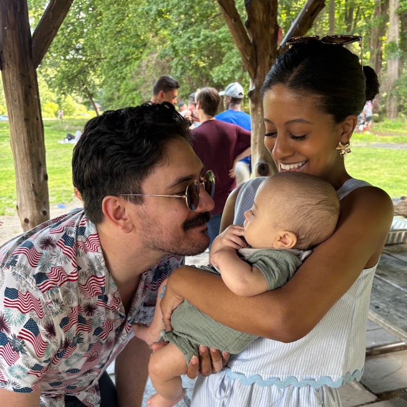 A dad is smiling with his face very close to his baby boy who is being held in the arms of his mom. The mother is smiling down at both dad and son. They are outside at a Grace Capital City Church holiday picnic, a church in Washington DC.
