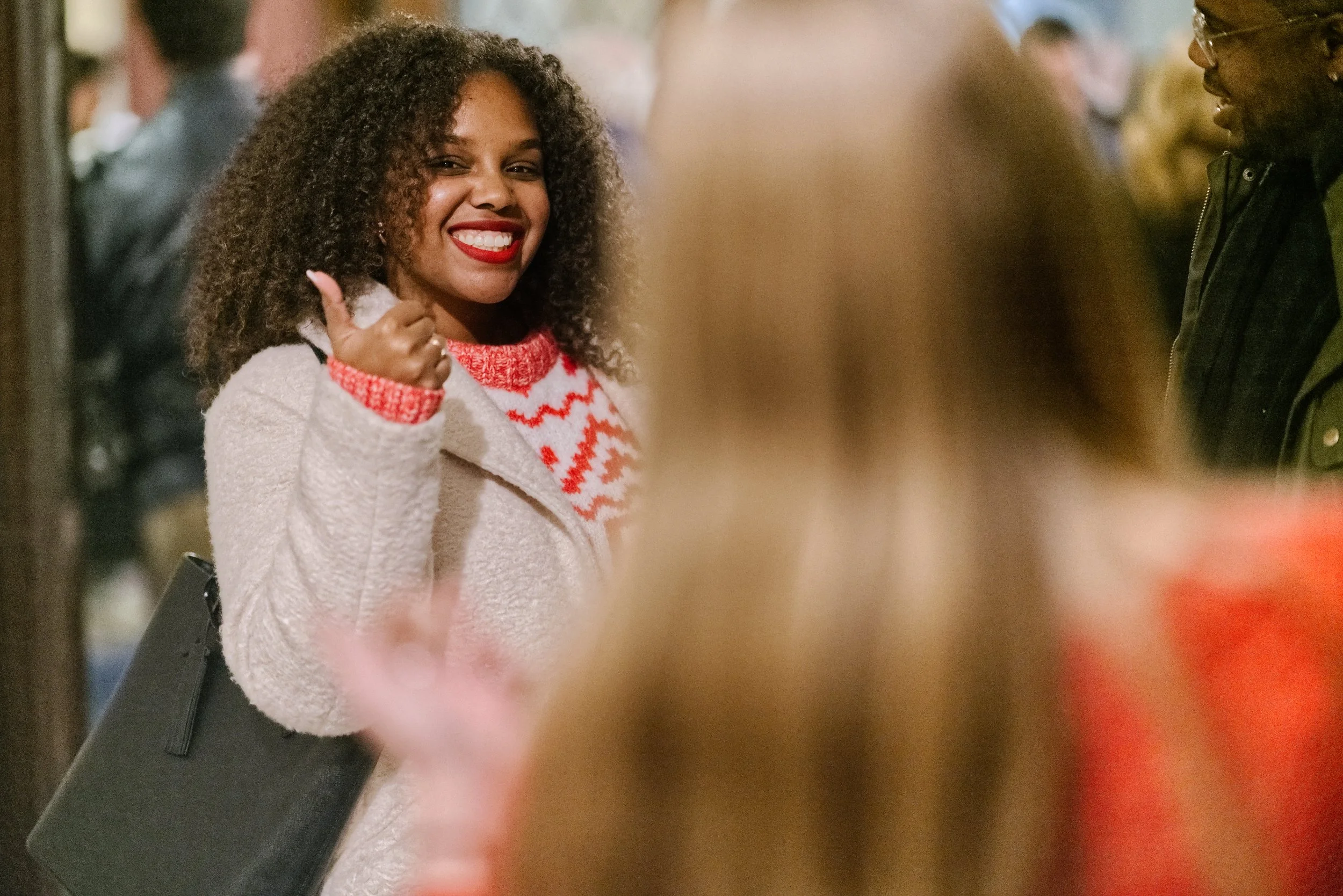 A smiling woman with curly hair wearing a white coat and red and white sweater giving a thumbs-up, standing with other people after a Grace Capital City church worship service in Washington, DC.