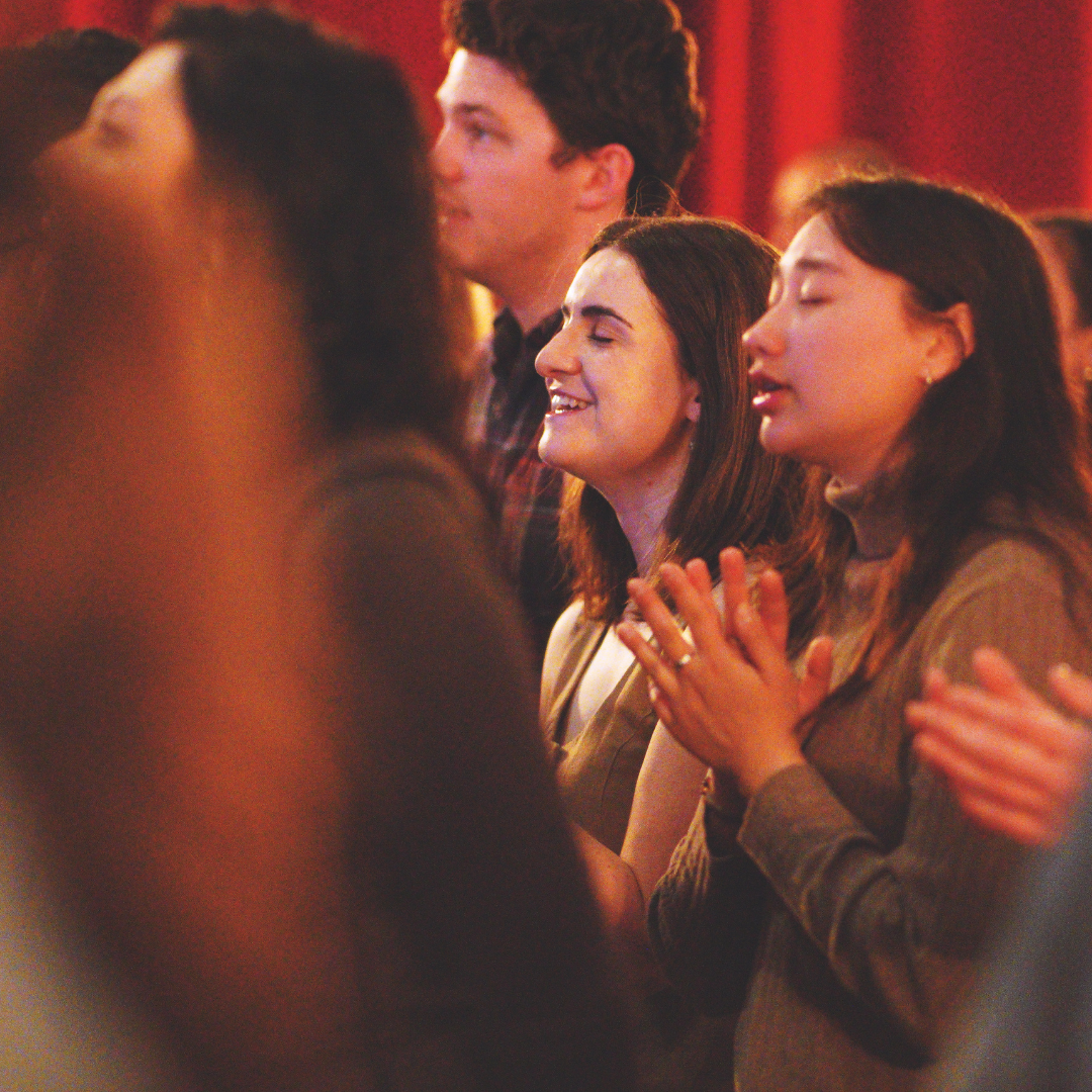 People worshipping during a Grace Capital City church service in Washington, DC. They are engaged and smiling with eyes closed, with dark-red curtains behind them.