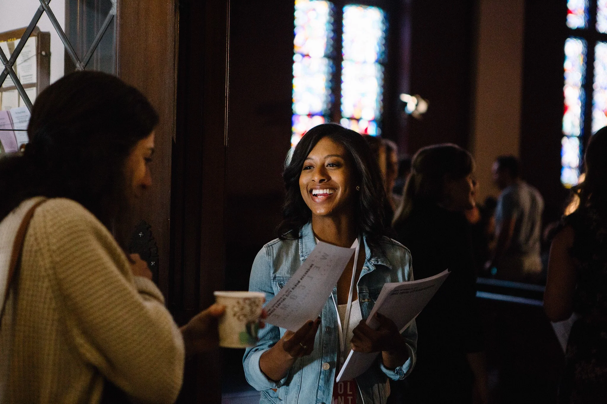 A woman in a denim jacket smiling and holding papers, talking to another woman holding a paper and a cup, inside a church with stained glass windows.