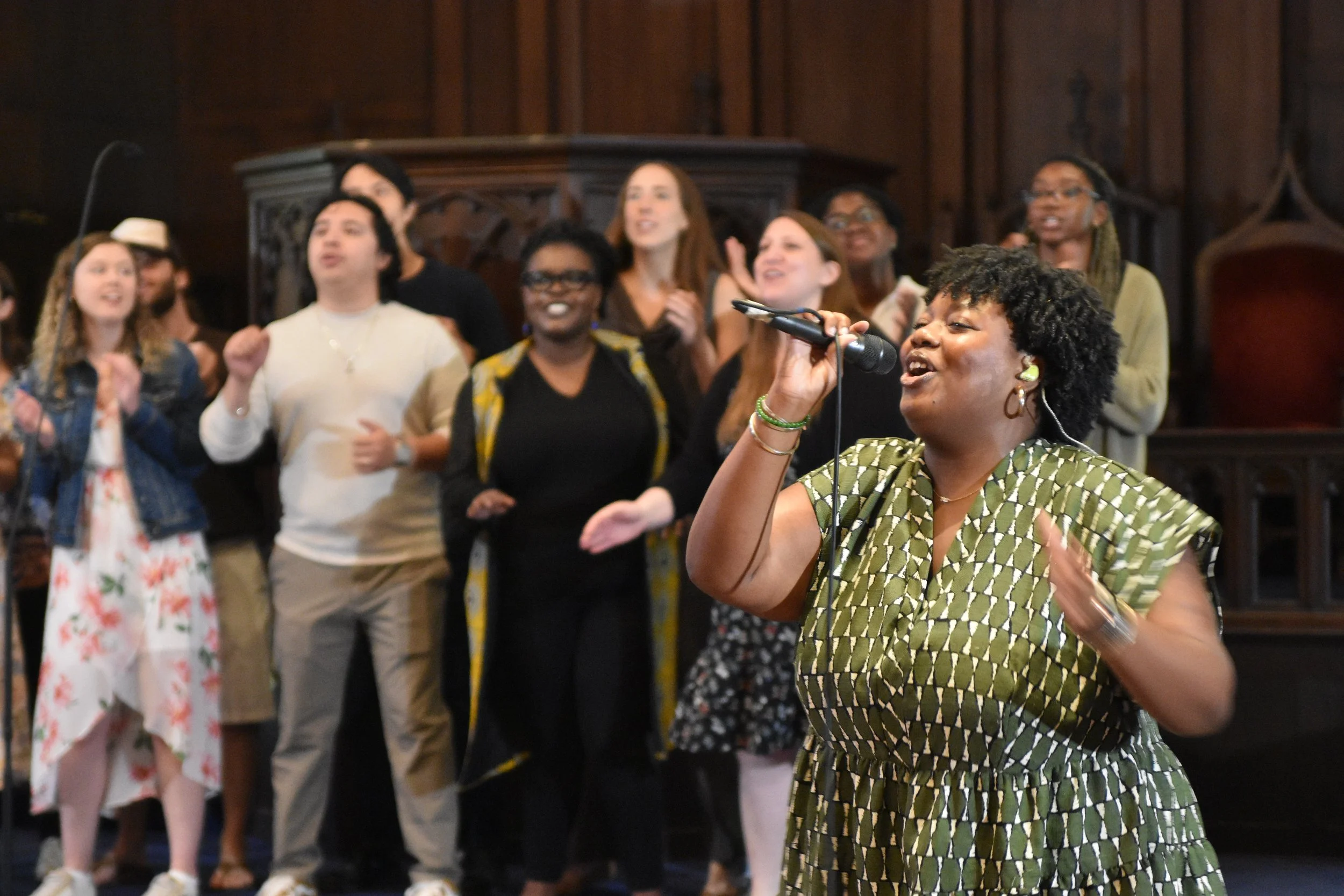 A woman worshipping, singing into a microphone surrounded by a diverse group of people in a gospel choir during a Grace Capital City church worship service in Washington, DC.