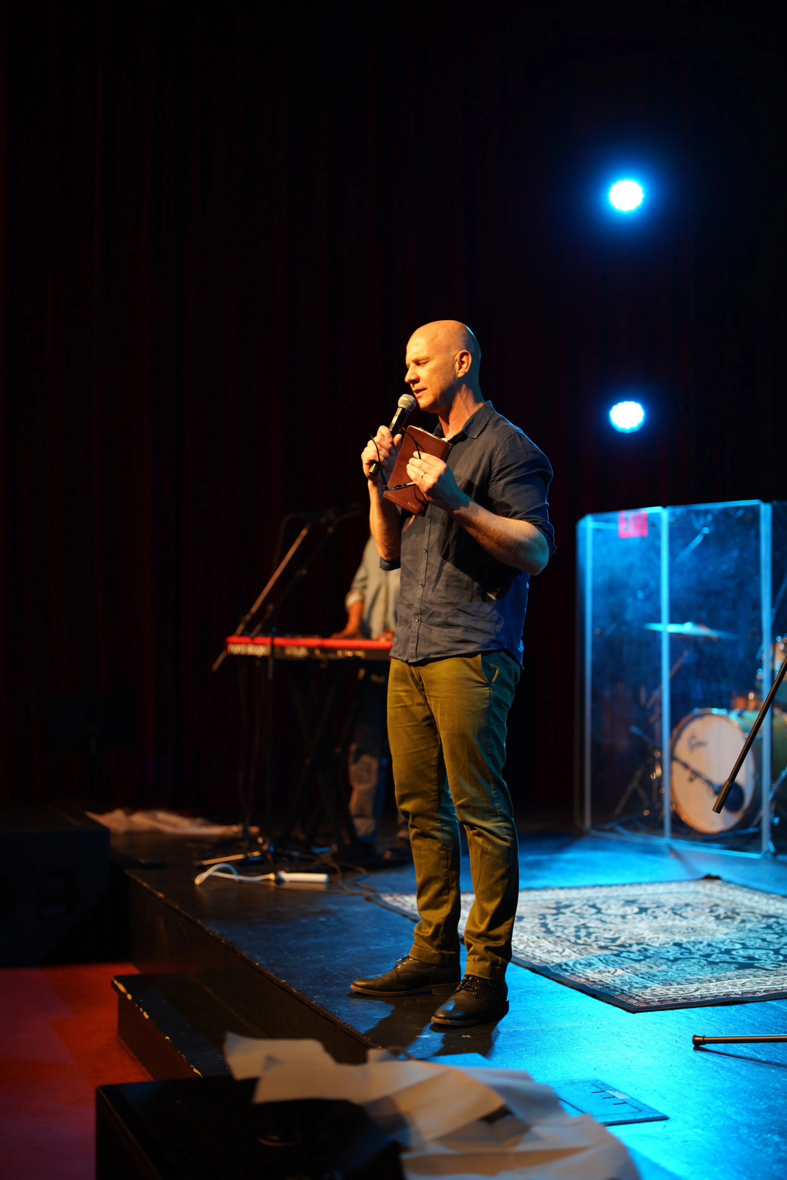 A man standing on a stage, holding a microphone and a notebook, speaking at a Grace Capital City church worship service. There is a band in the background with stage lights shining down.