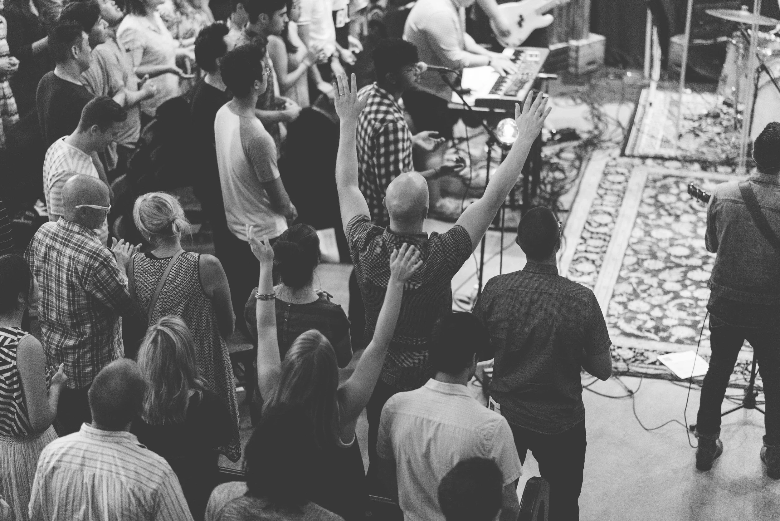 A crowd of people at a Grace Capital City church worship service, some with hands raised, gathered around musicians playing instruments and a microphone on a patterned rug.