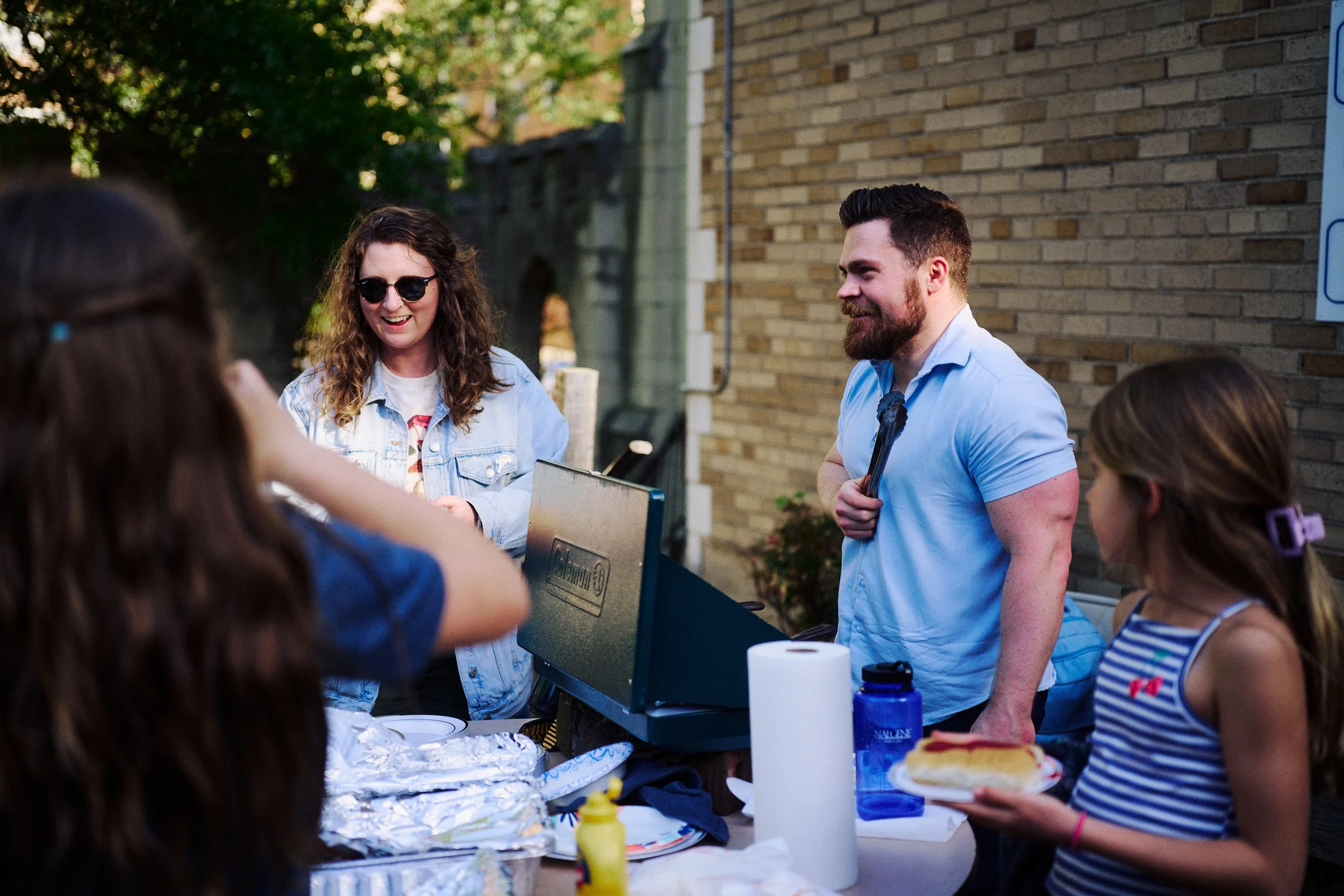A group of friends at a cookout in Washington, DC for Grace Capital City church, with a man holding tongs, a woman with sunglasses smiling, and a girl holding a plate of food, around a table with food and supplies.