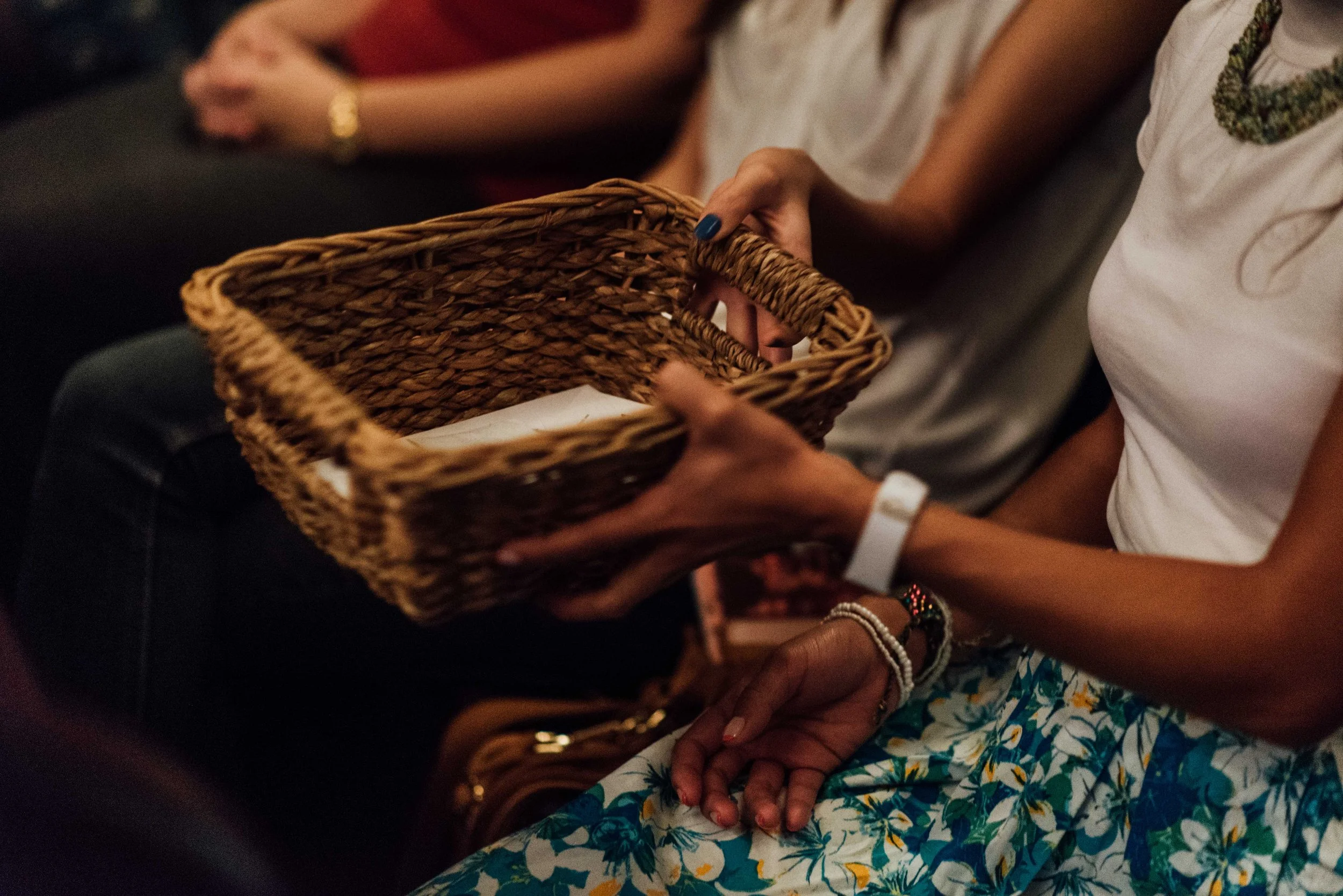 A person passing a woven basket during a service at Grace Capital City church. The person is sitting next to another person with hands clasped on their lap, wearing a white t-shirt, floral skirt, and colorful jewelry.