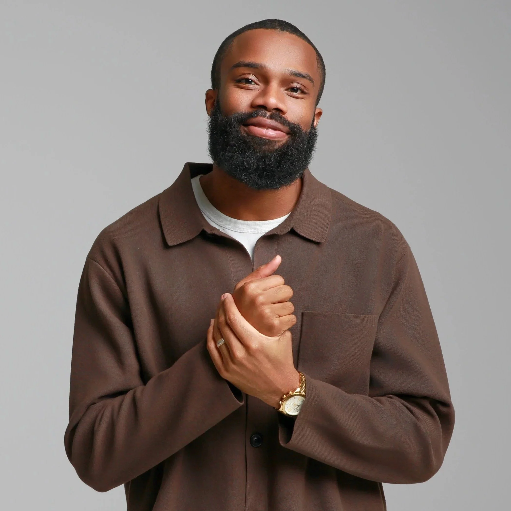A smiling Black man with a beard, wearing a brown jacket, white shirt, watch, and a ring, is standing against a plain light gray background. He serves as a Pastor at Grace Capital City, a church in Washington, DC.