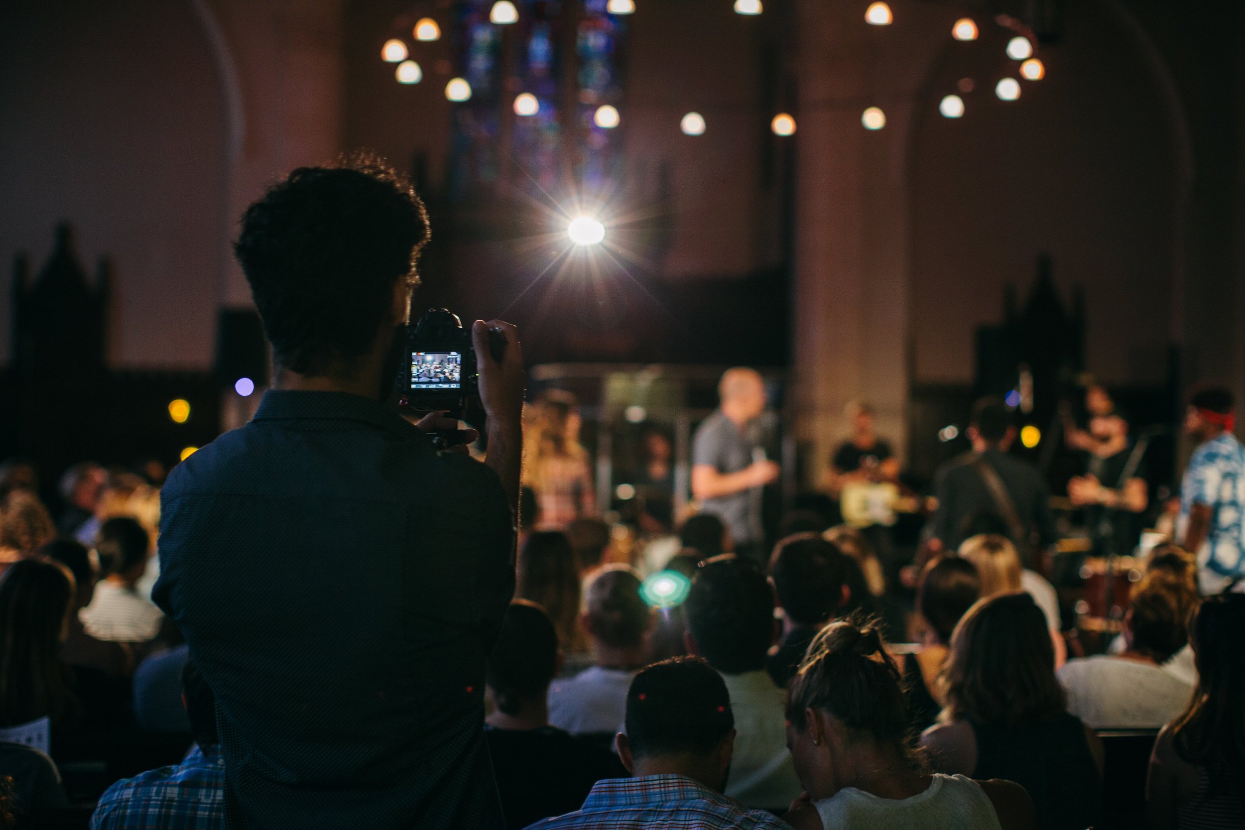 A person in the foreground is taking a photo or recording a video of a band performing on stage inside a church with stained glass windows and hanging lights.