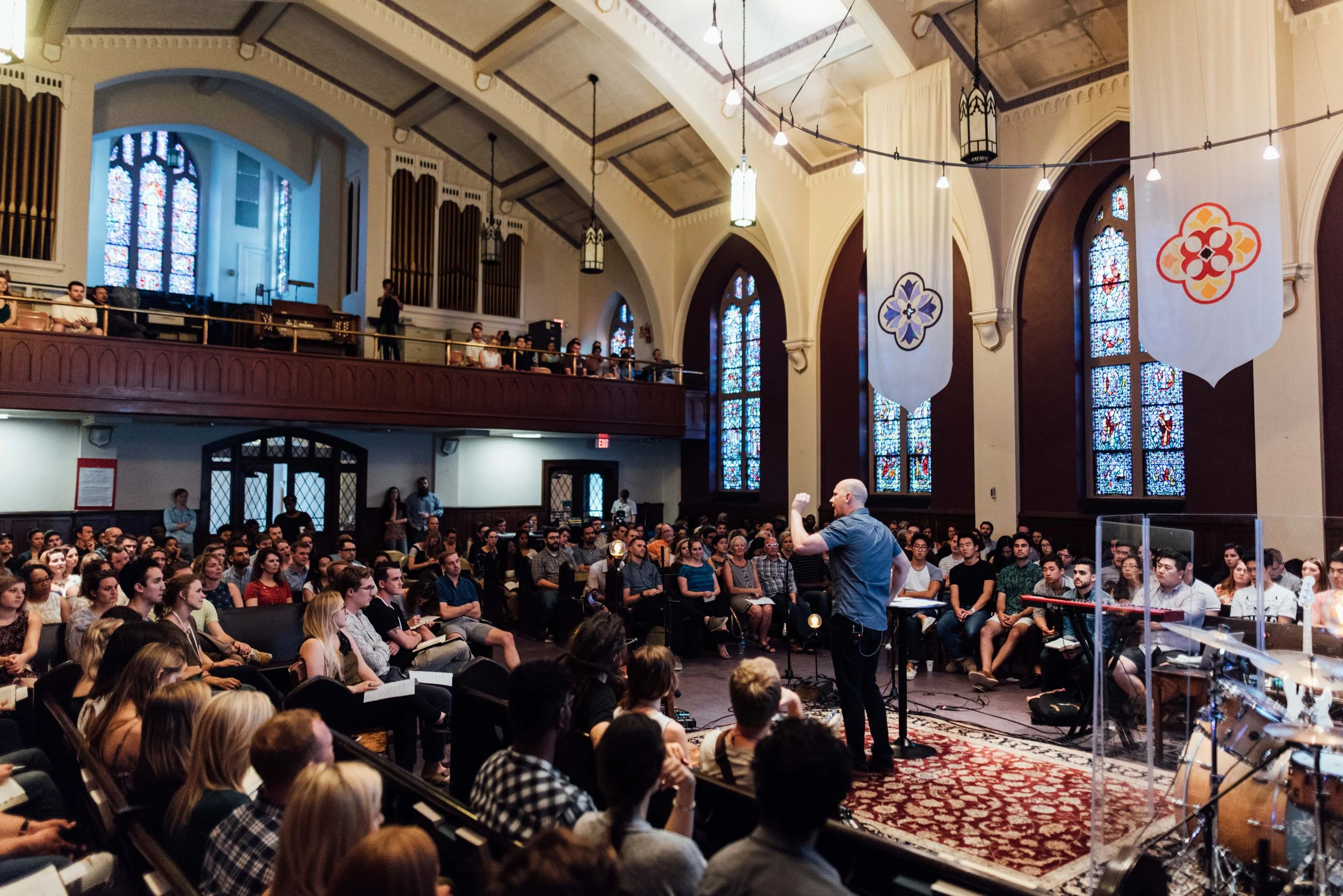 A large group of people gathered for a Grace Capital City church worship service in Washington, DC with stained glass windows and banners hanging from the ceiling. A man is speaking in front of them.