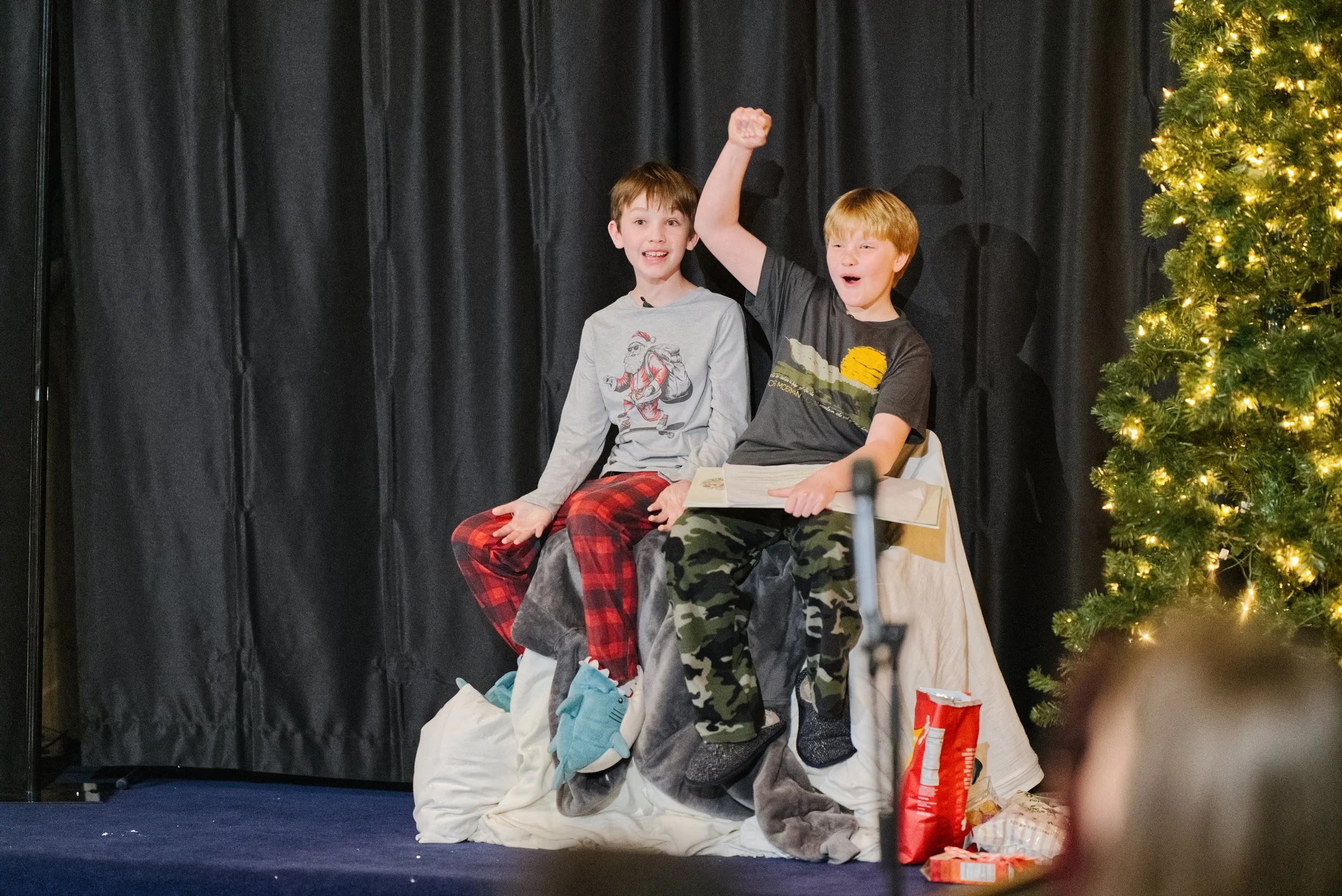 Two boys in pajamas are sitting on a decorated stage with a Christmas tree to their right during a Grace Capital City church Christmas service for Children's Ministry.