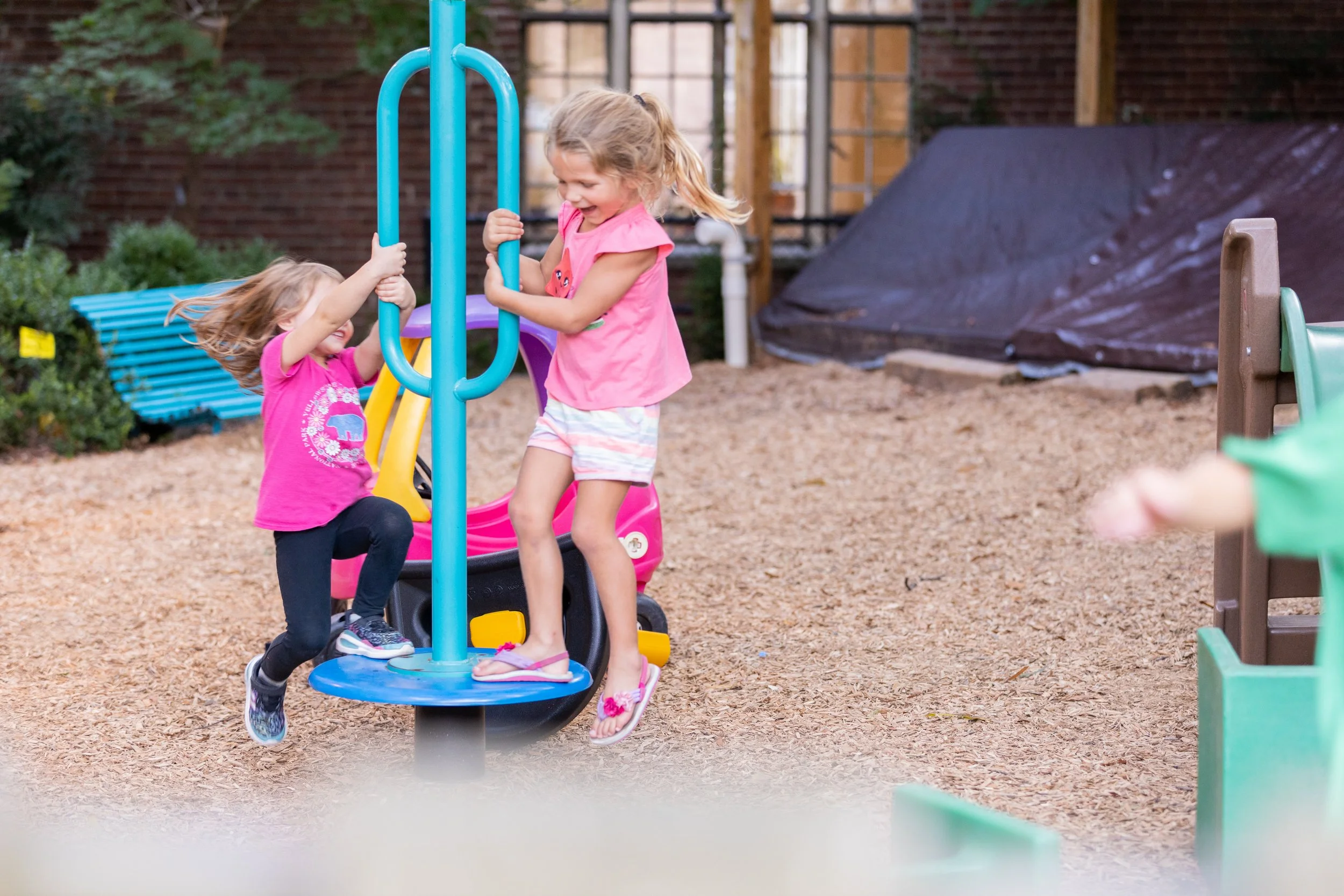 Two young girls are playing on a circular merry-go-round in a playground, holding onto the blue handles and smiling after a Grace Capital City church worship service in Washington, DC.