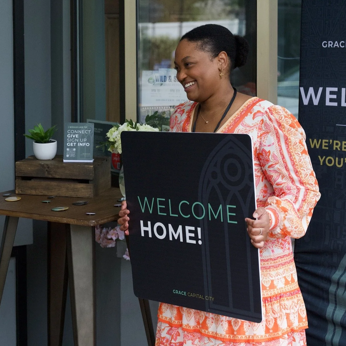 A smiling woman holding a sign that says 'Welcome Home!' while greeting guests before a Grace Capital City church worship service in Washington, DC.