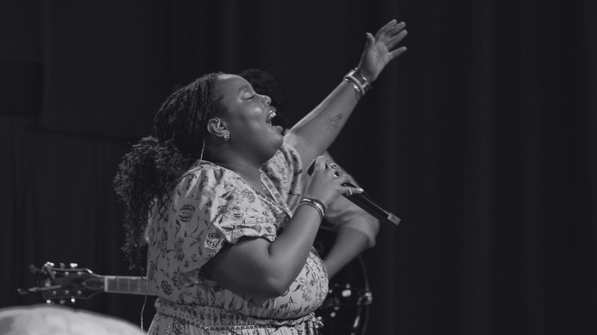 A woman singing passionately with her eyes closed at a Grace Capital City church worship service. She is holding a microphone in one hand and raising her other hand, on stage with a dark background, wearing a patterned dress and bracelets.