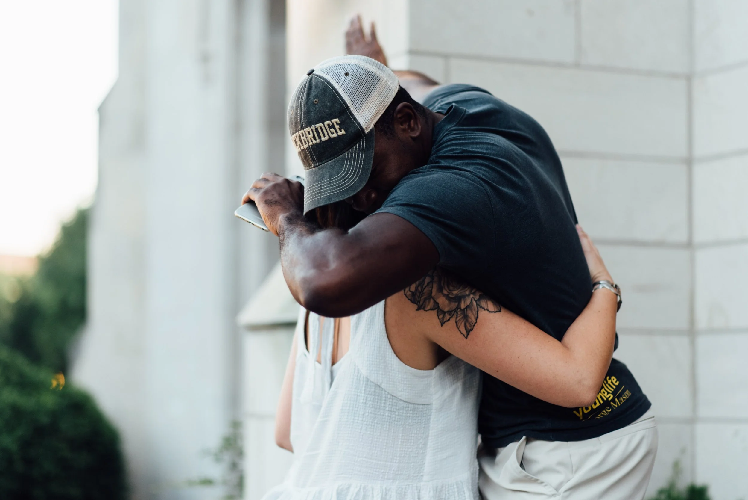 Two people hugging outside Grace Capital City church in Washington, DC, one wearing a baseball cap and the other a white dress.