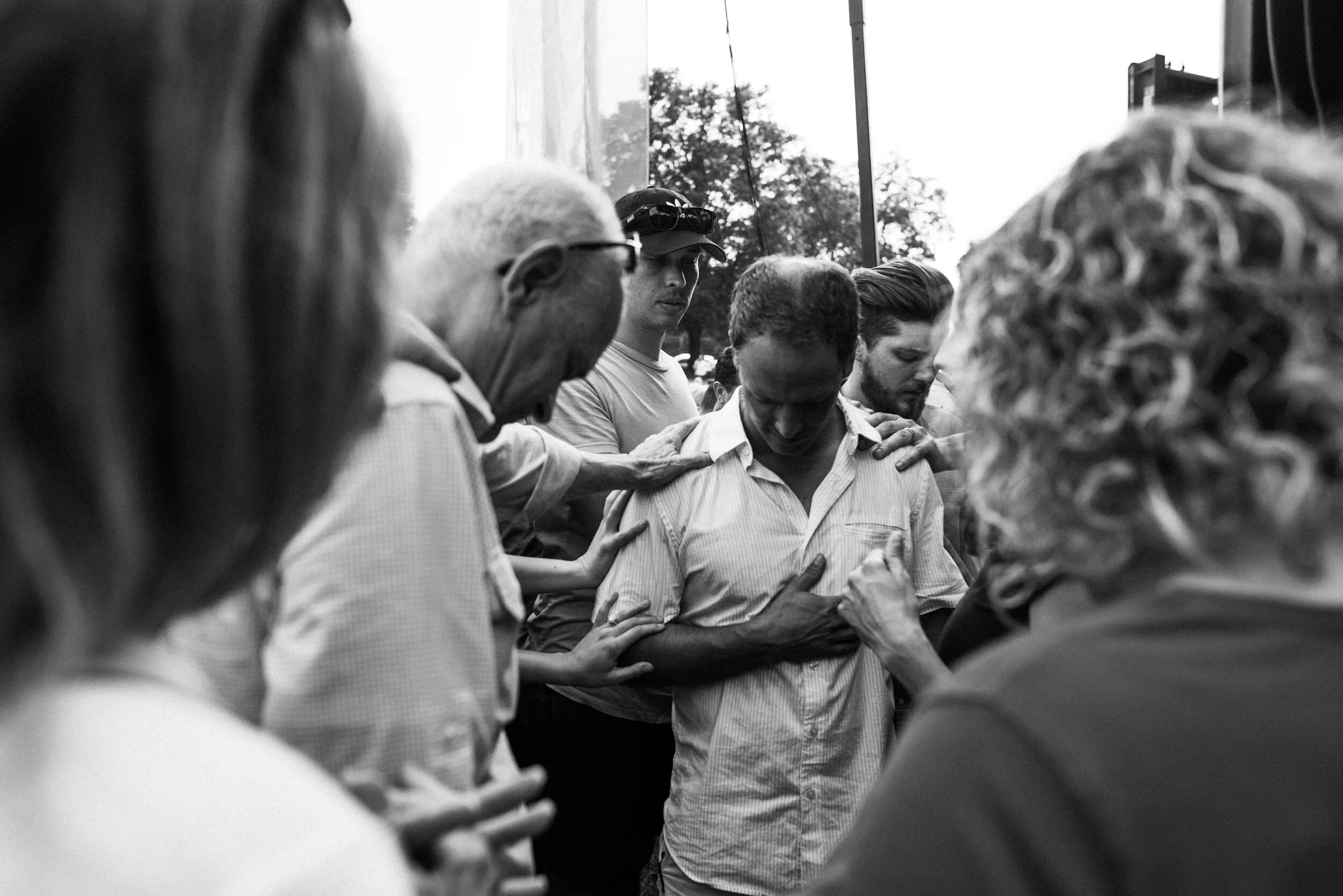 A group of people, with heads bowed and hands placed on each other's shoulders, praying before a Grace Capital City church worship service in Washington, DC.