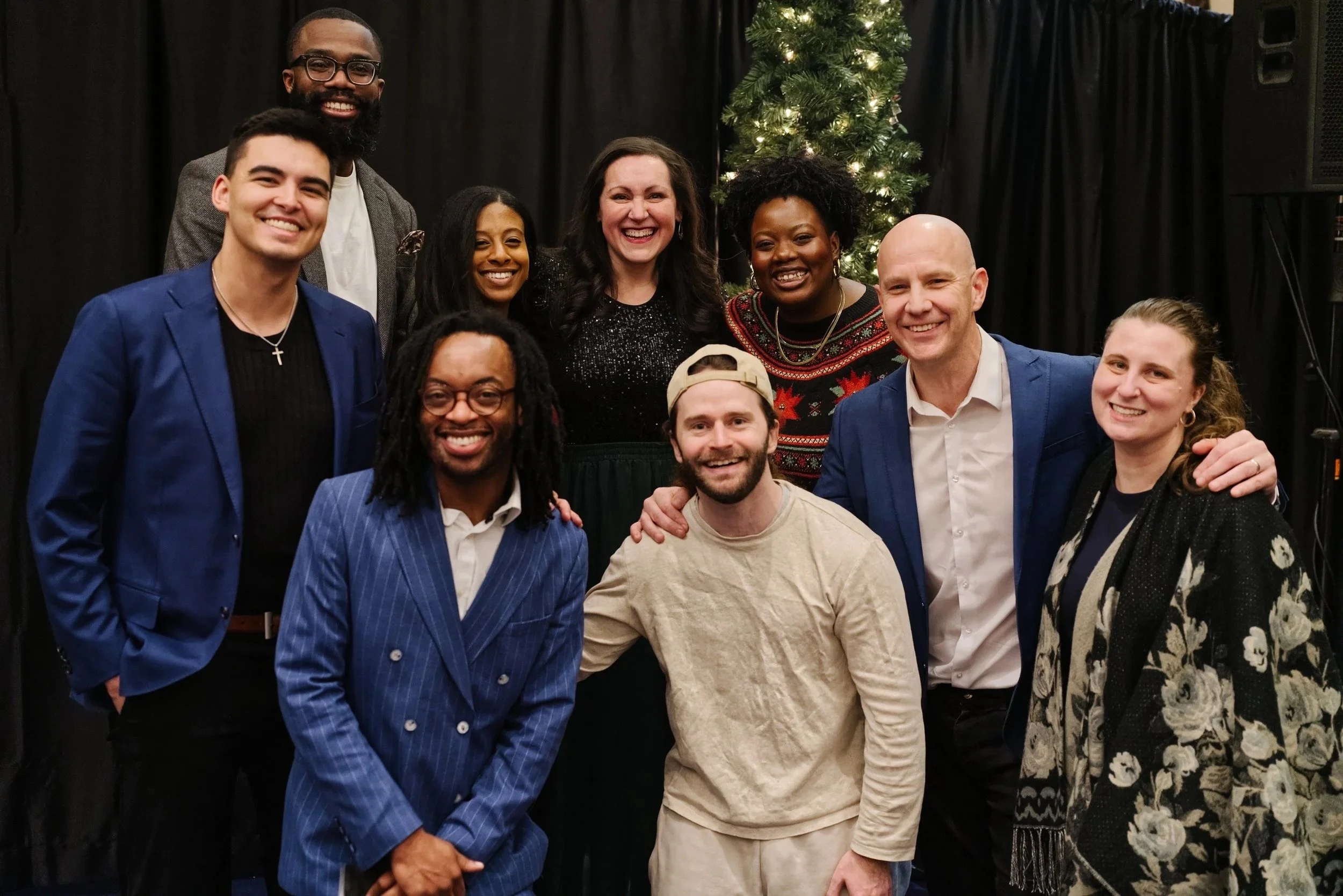 The staff of Grace Capital City, a group of ten diverse people, smiling in front of a Christmas tree, dressed casually and formally, celebrating together after a Grace Capital City church worship service in Washington, DC.