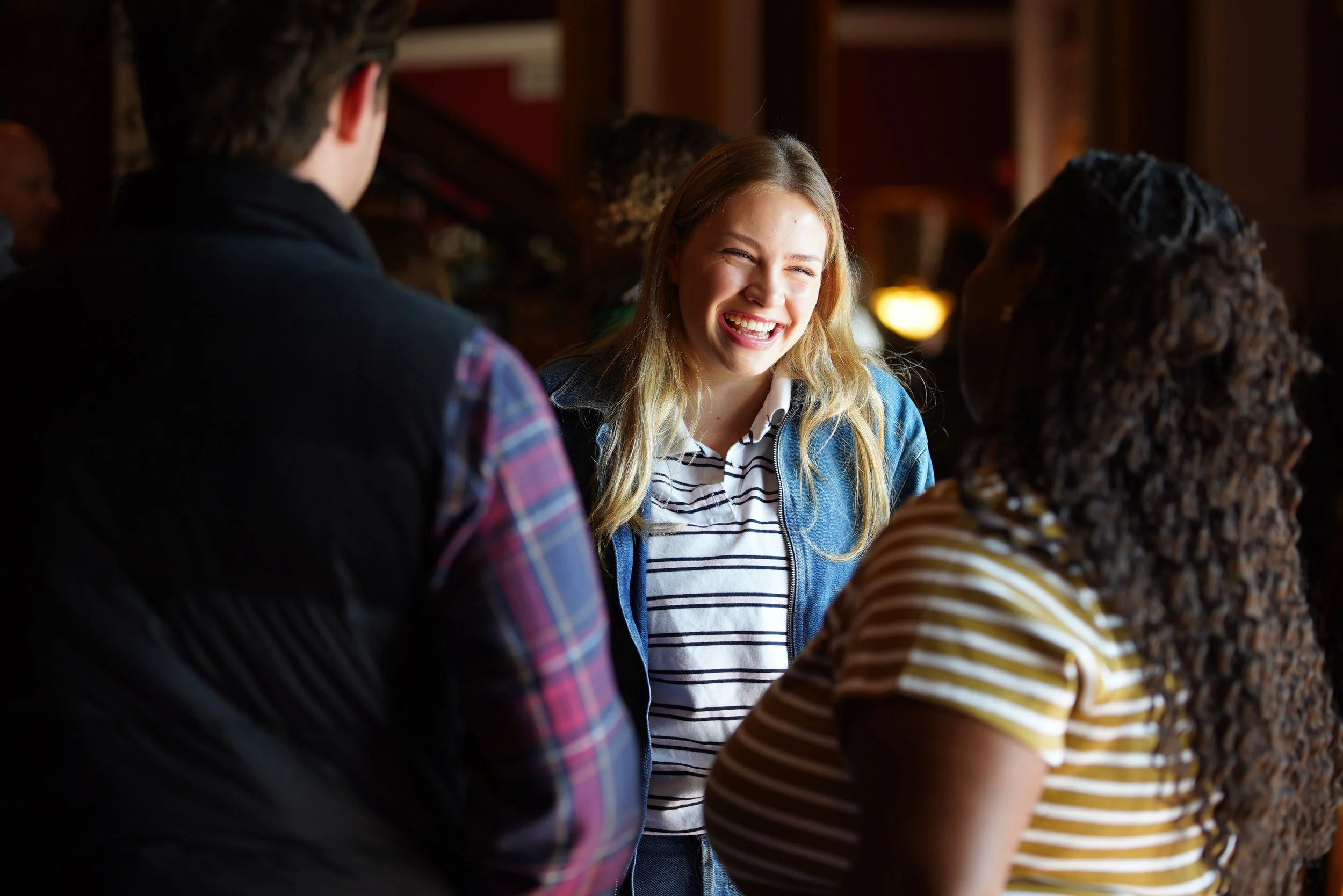 A group of  friends, two women and one man, are having a conversation after a worship service at Grace Capital City church. Centered is a blonde-haired woman in a denim jacket and striped-shirt. The back of the other two people are in front of her.