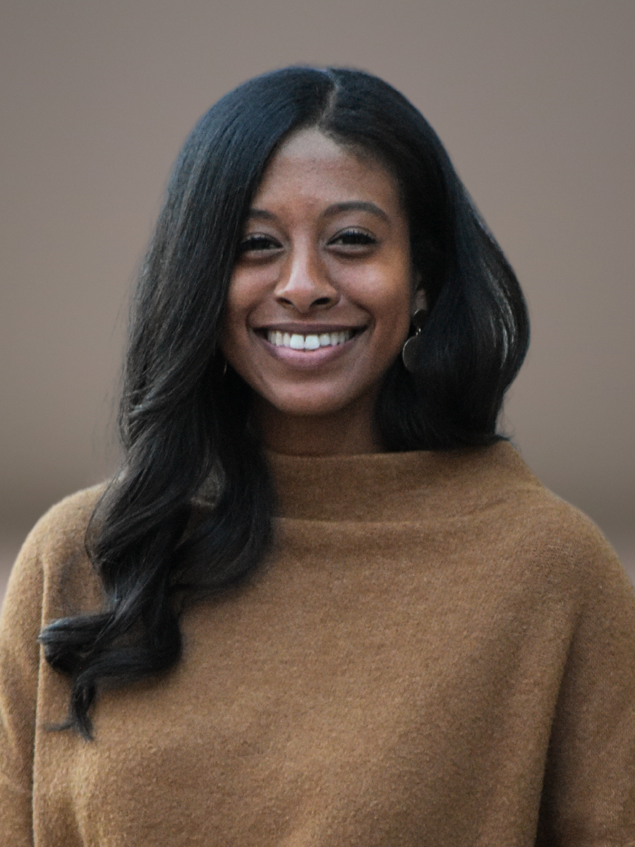 A smiling dark-skinned woman with long black hair, wearing a brown sweater, stands against a blurred background. She serves on staff at Grace Capital City, a church in Washington, DC.