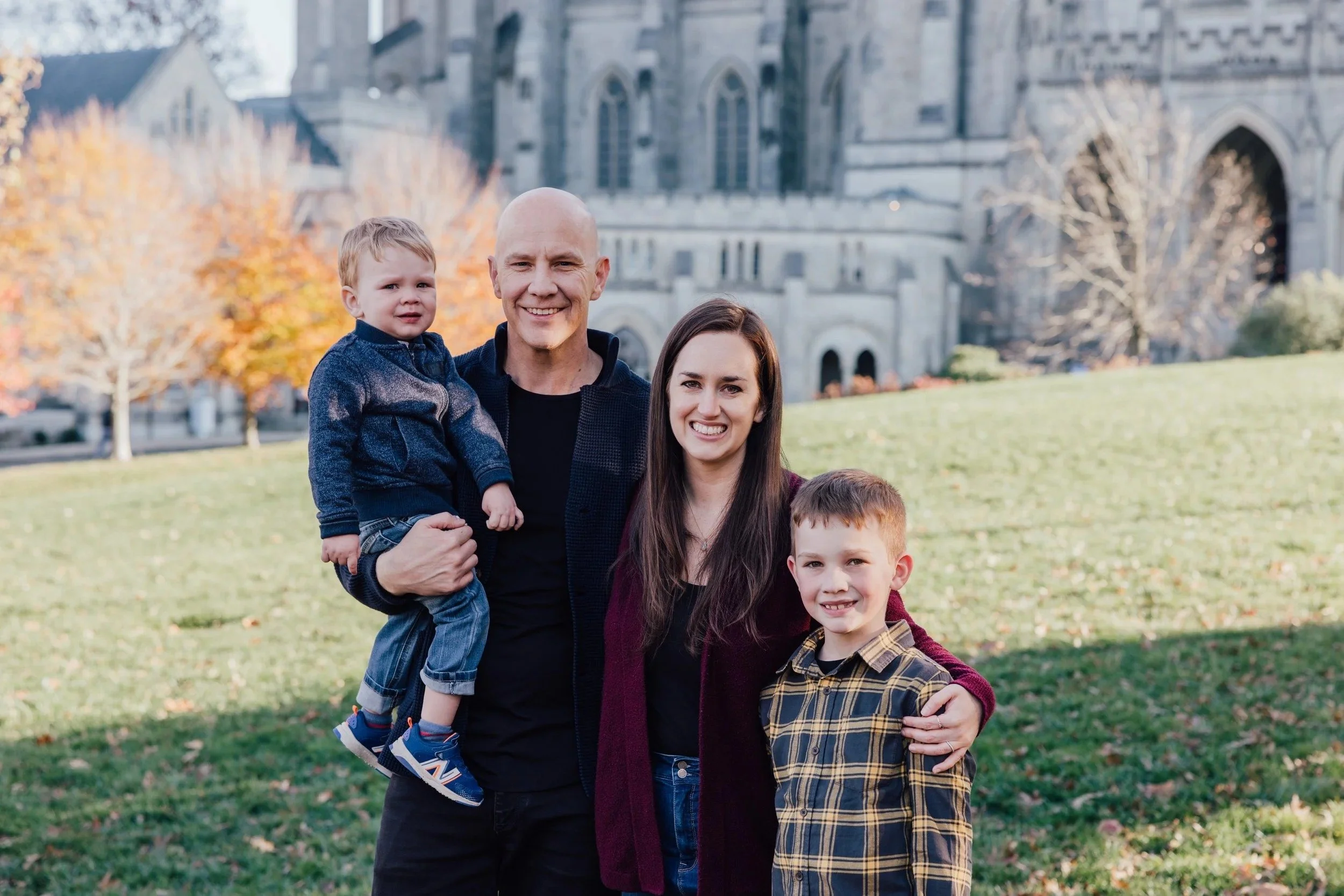 A happy family of four standing outdoors on a sunny fall day in front of a historic stone church, with autumn trees in the background that helped start Grace Capital City, a church in Washington, DC.