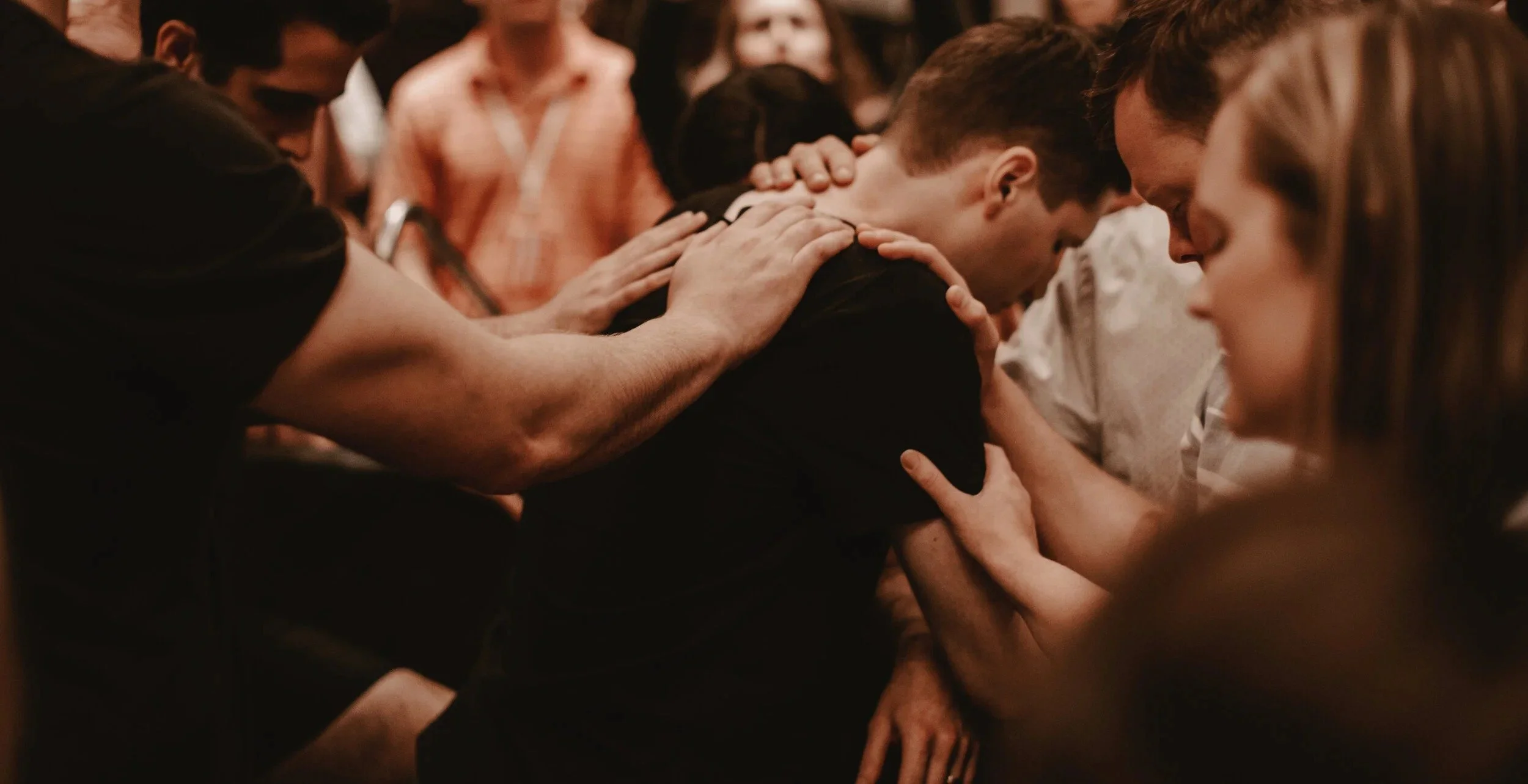 Group of people praying or mourning closely together with heads bowed and hands on each other's shoulders and back.