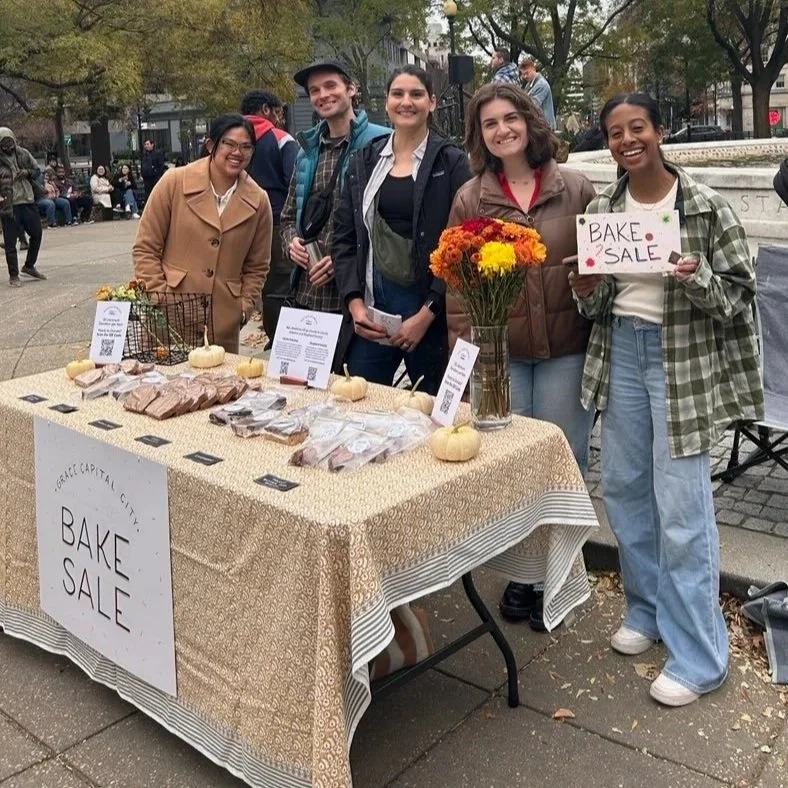 Group of five young people with Grace Capital City church standing behind a table at a bake sale in a Washington, DC park, with baked goods and a sign that has "Bake Sale" on it, with trees and people in the background.