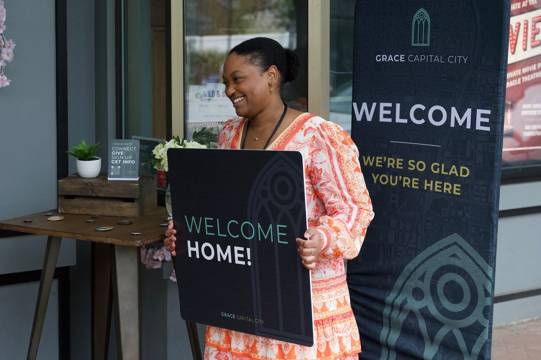 A woman standing at a table outdoors and smiling at a Grace Capital City church service, holding a welcome sign that says "Welcome Home!" behind her, a large standing banner reads "Welcome, We're so glad you're here."