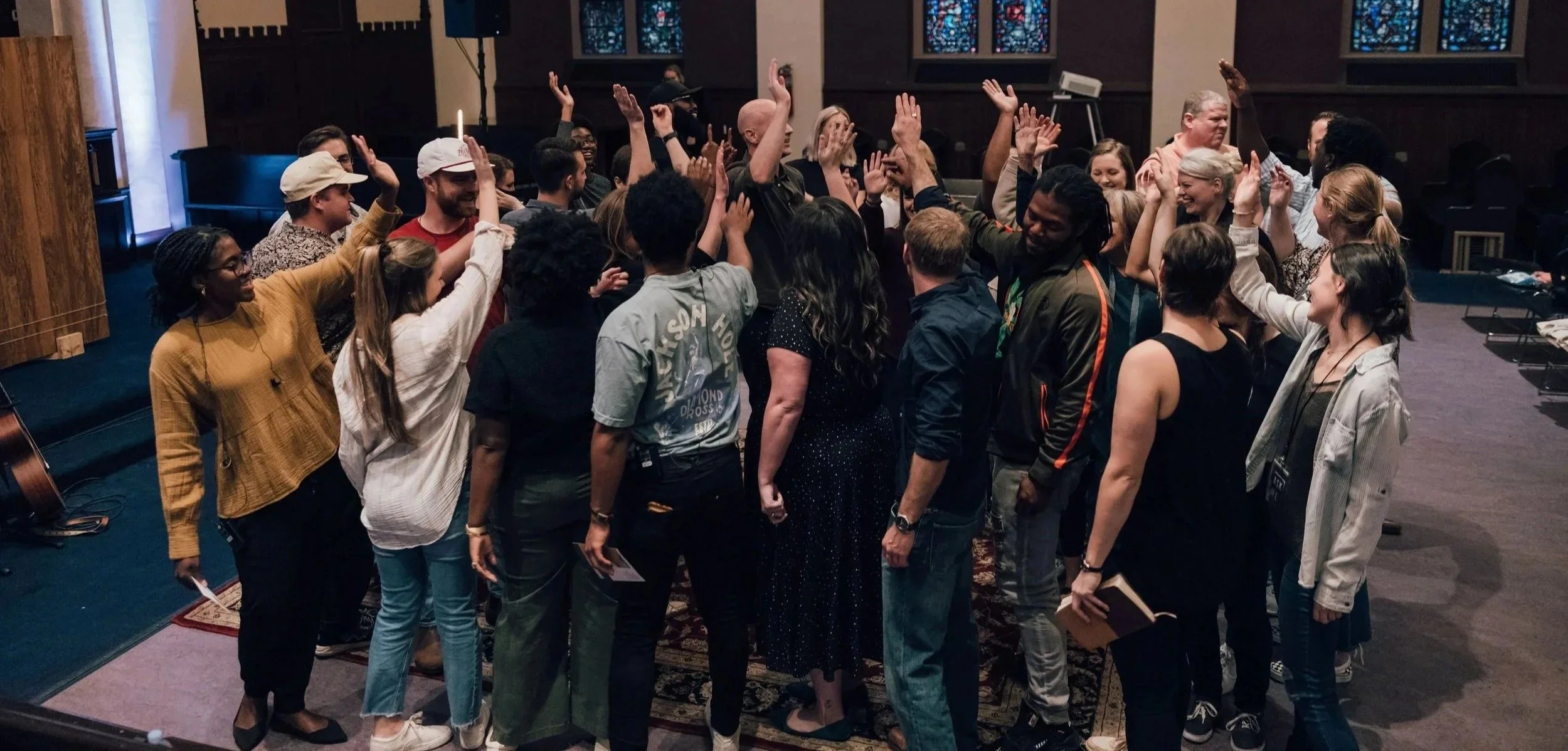A group of diverse people standing in a circle with raised hands, celebrating before a Grace Capital City church worship service in Washington, DC with dark walls and stained glass windows.