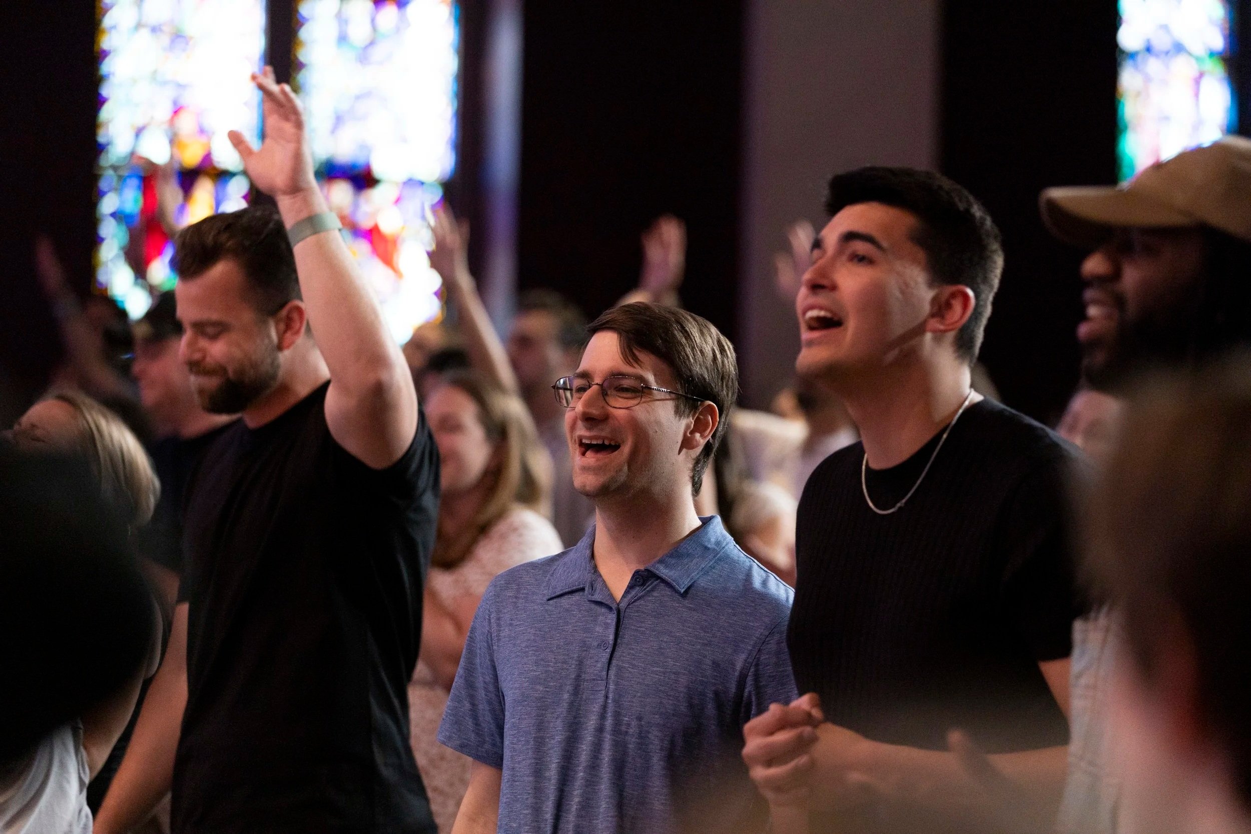 A group of people at a Grace Capital City church worship service, singing and raising their hands, with stained glass windows in the background.