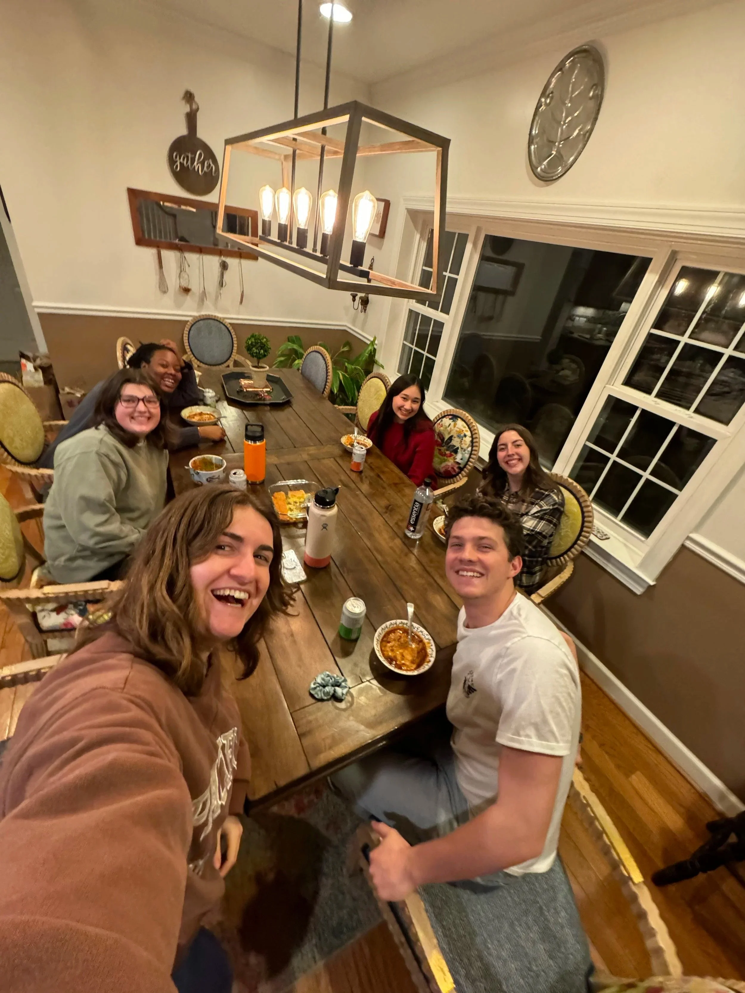 A group of six young adults sitting around a dining table, smiling and enjoying a meal together in a cozy dining room with large windows and decorative chairs during a gathering for College Ministry at Grace Capital City, a church in Washington, DC.