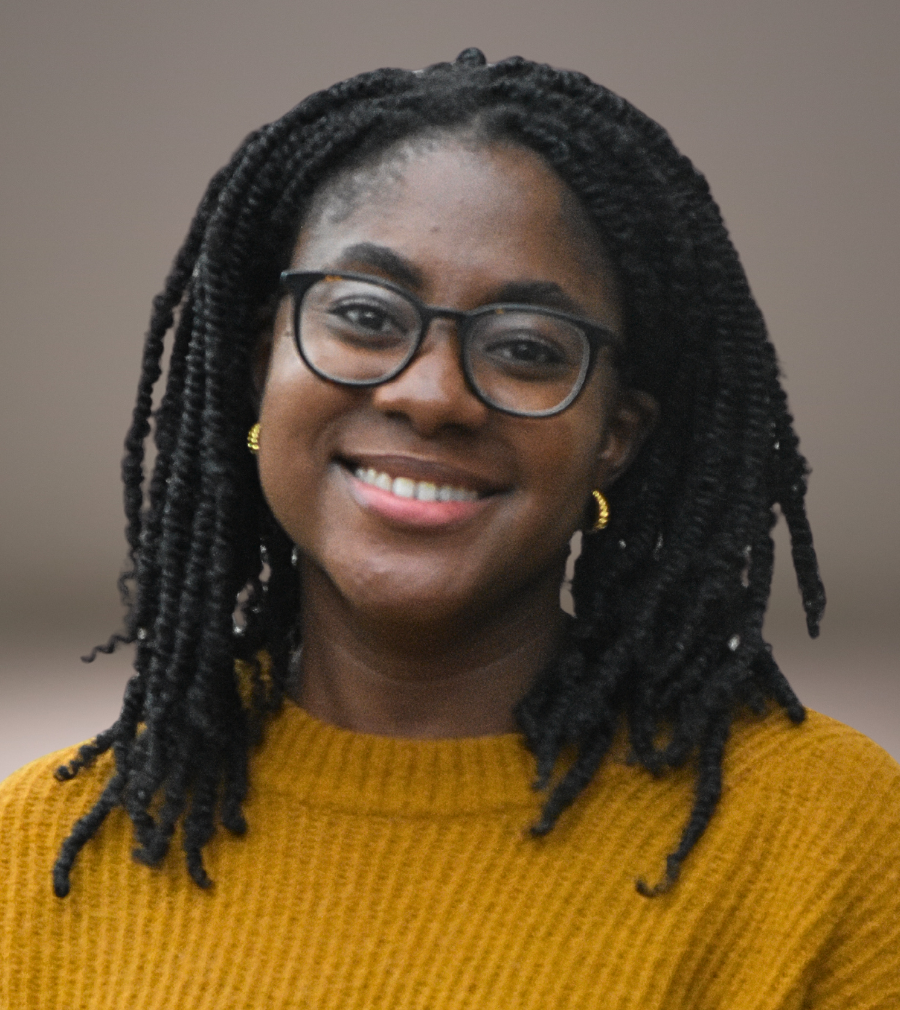 A woman with dark braided hair, glasses, and gold earrings smiling in a mustard sweater against a plain background.