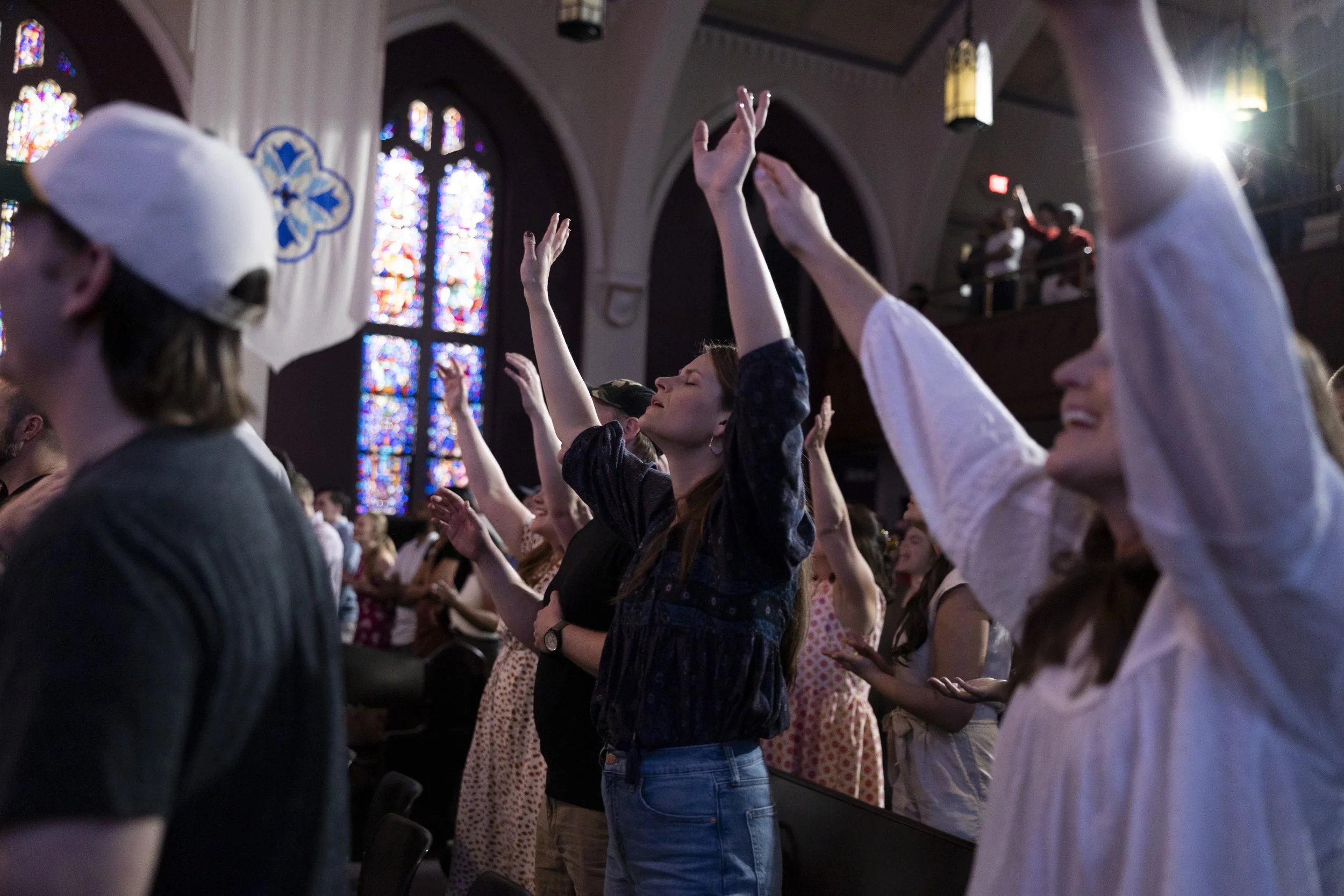 People inside a church with stained glass windows, raising their hands in worship during a Grace Capital City church worship service in Washington, DC.