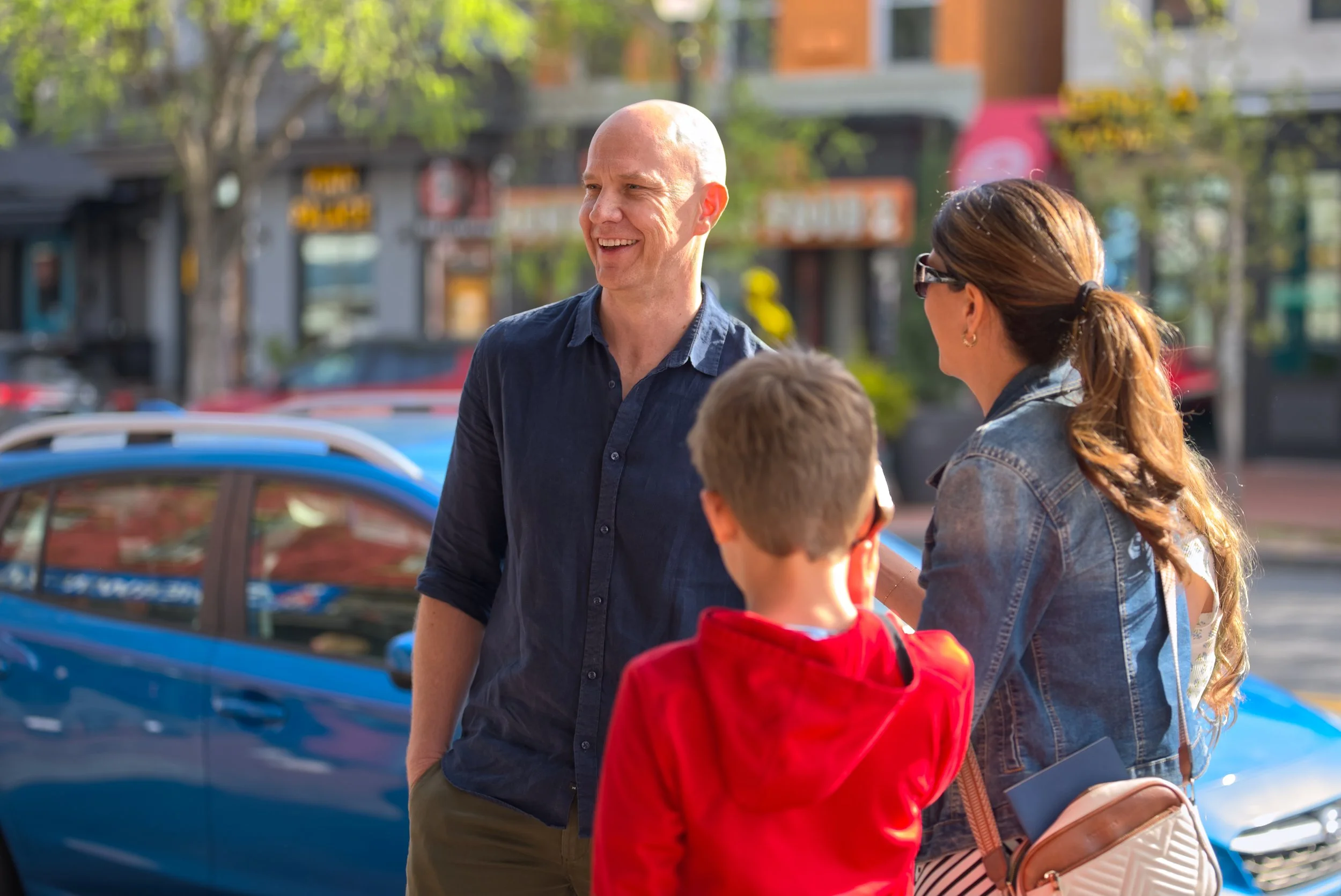 A man and a woman are talking to a child on a city sidewalk during the day. The man is bald and smiling, wearing a dark blue shirt. The woman has long hair tied back, sunglasses, a denim jacket, and is holding a small purse. A blue car is parked behind them, and trees and storefronts are visible in the background.