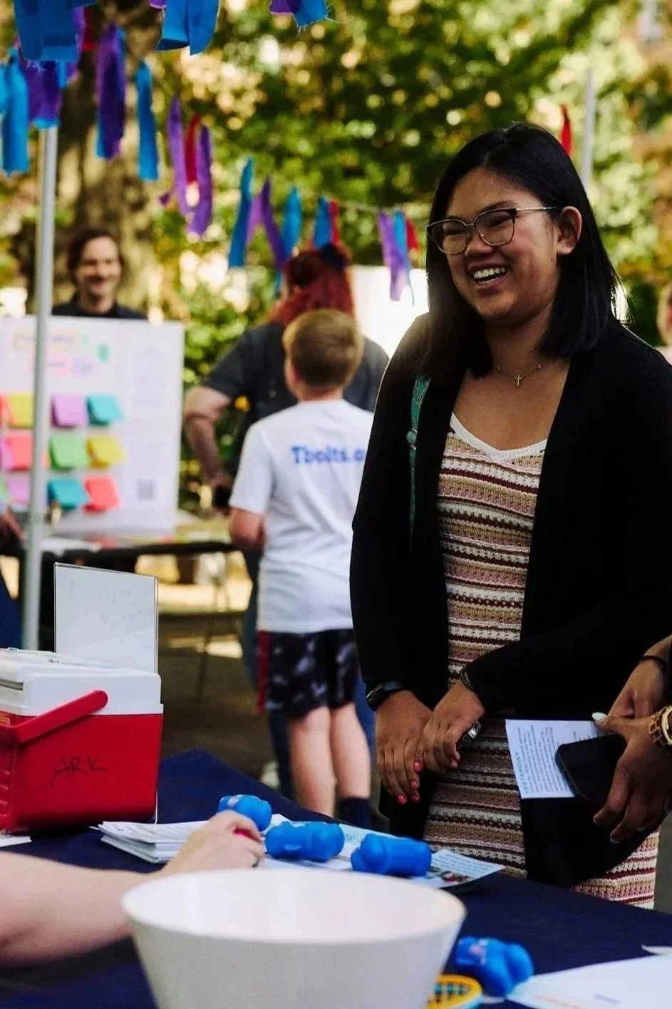 People at an outdoor event for Grace Capital City church in Washington, DC, with colorful decorations, a woman smiling in the foreground, a table with items, and children and adults in the background.