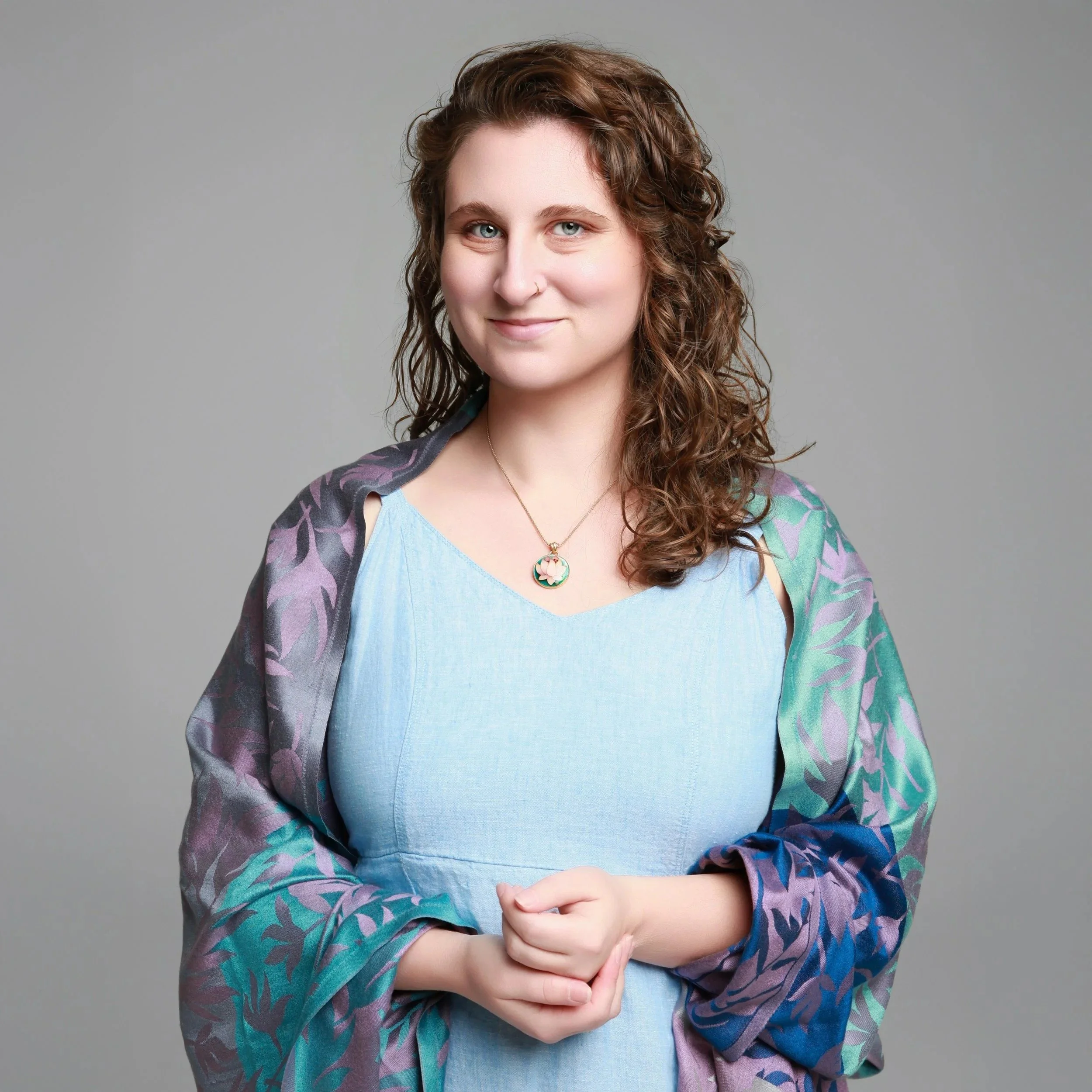A woman with shoulder-length curly brown hair, blue eyes, and fair skin, smiling slightly, wearing a light blue dress with a multicolored patterned shawl and a gold necklace with a colorful pendant, standing against a plain gray background.
