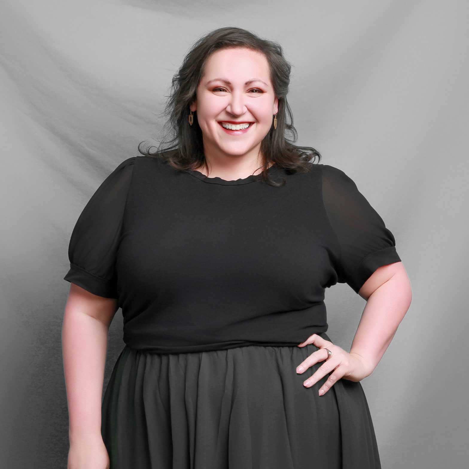 A white woman with shoulder-length dark hair, smiling, wearing a black top with puffed sleeves and a black skirt, standing against a plain gray background. She serves as the Associate Pastor at Grace Capital City, a church in Washington, DC.