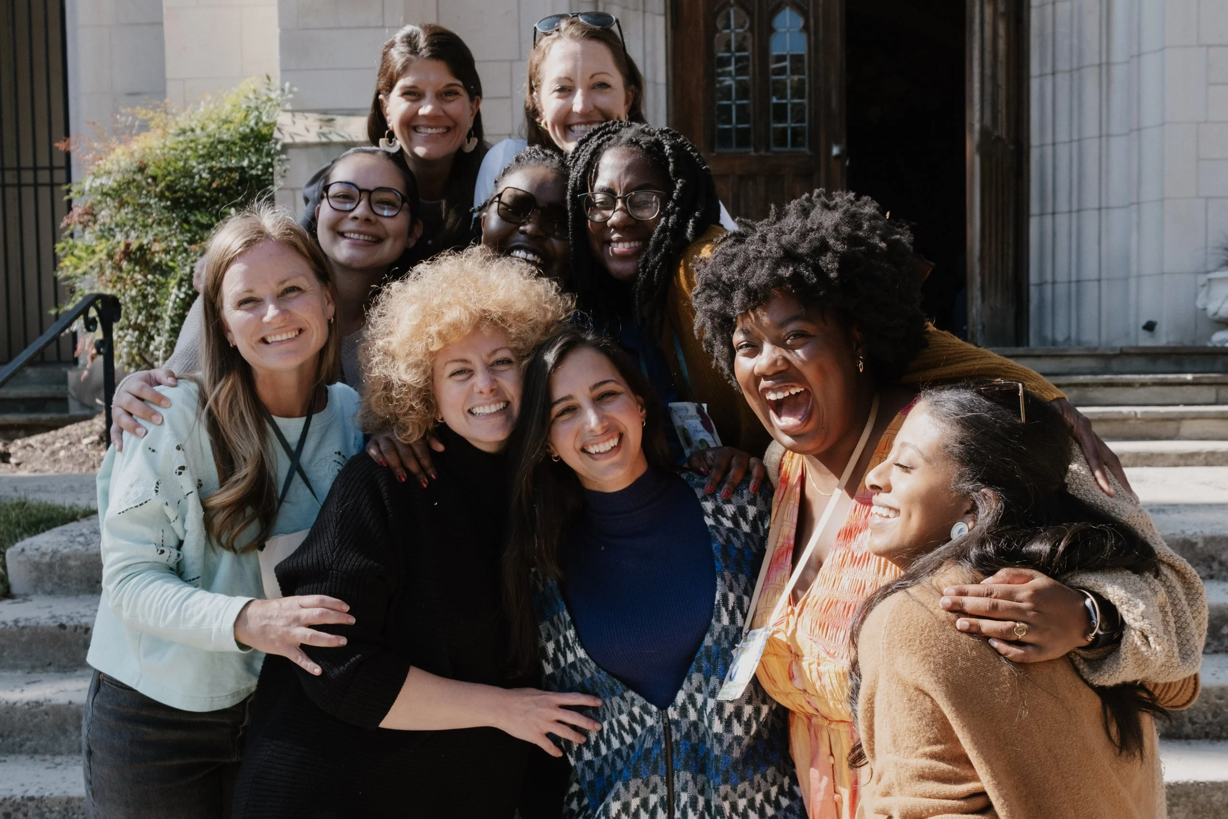 A group of diverse women outdoors on the steps of a building, smiling and embracing each other in a joyful moment after a GCC Women's gathering for Grace Capital City church in Washington, DC.