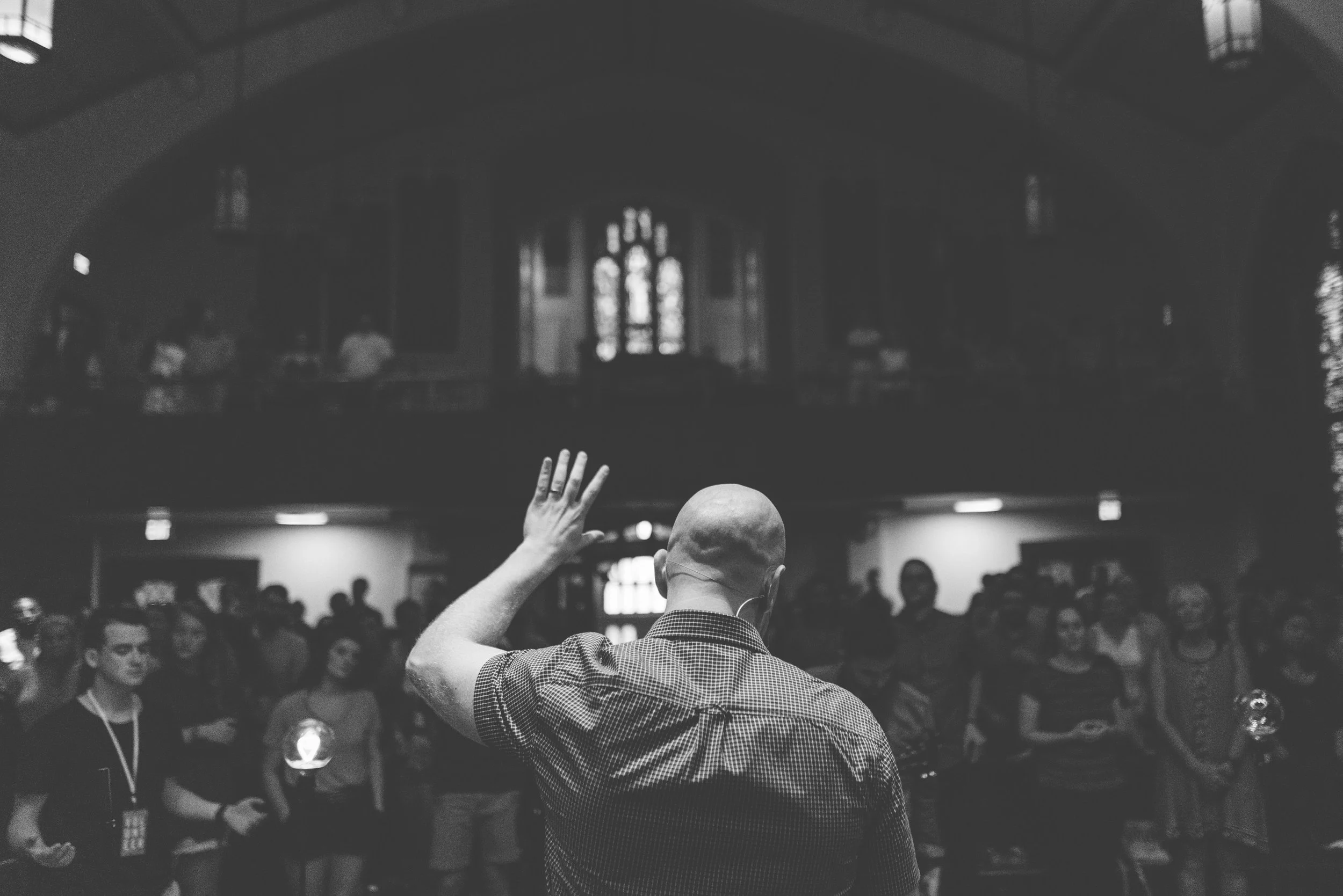A man with no hair on his head and wearing checkered blue, short-sleeved shirt raising his hand in front of a group of people in a churc during a gathering or service, at Grace Capital City church.