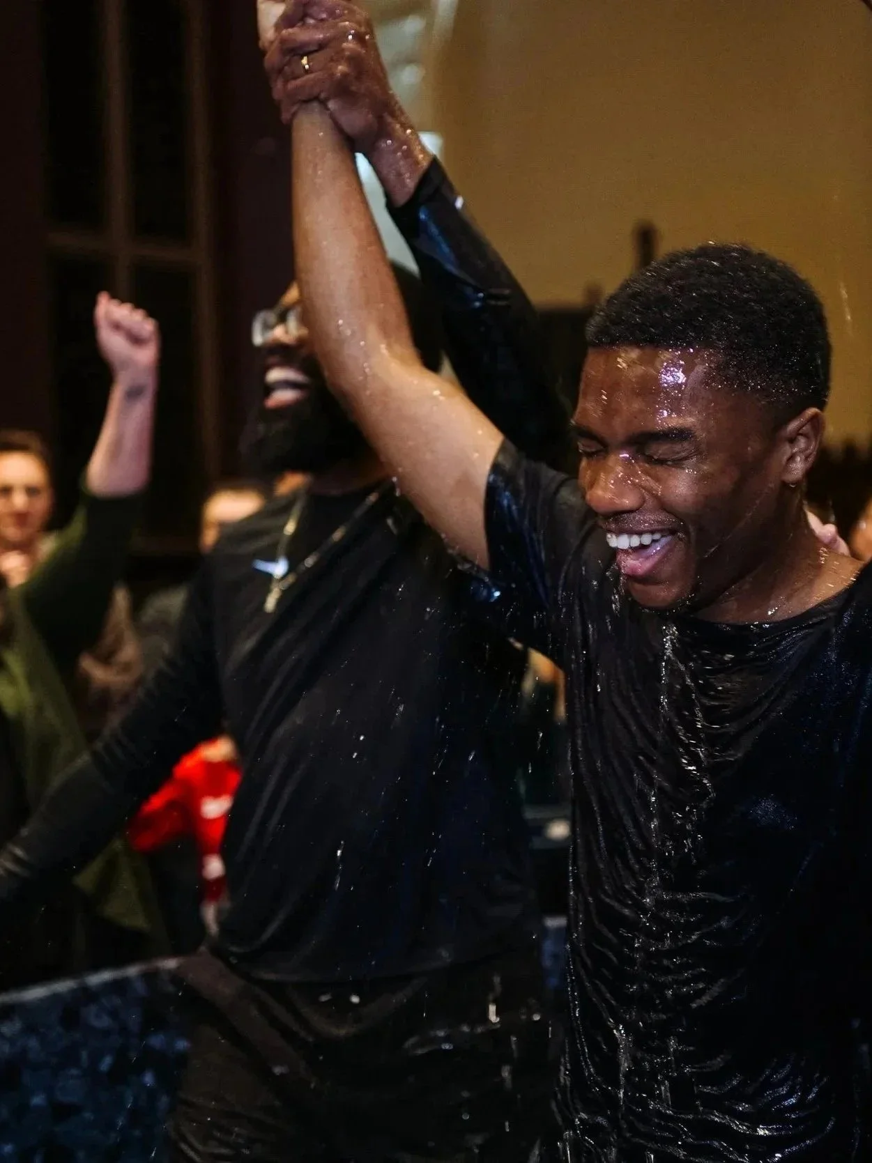 A photo of a young man getting baptized during a Grace Capital City church worship service.  The young man is in the foreground smiling with eyes closed, wearing a black shirt, water on his face and body, while others are visible in the background.