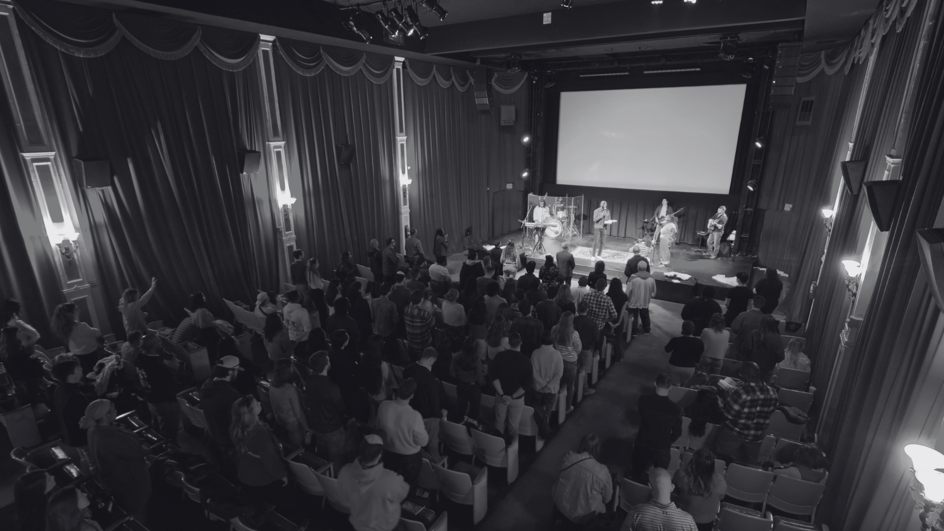Crowd of people attending a Grace Capital City church worship service in a theater with a large screen behind a worship band, and with curtains and decorative wall lighting.