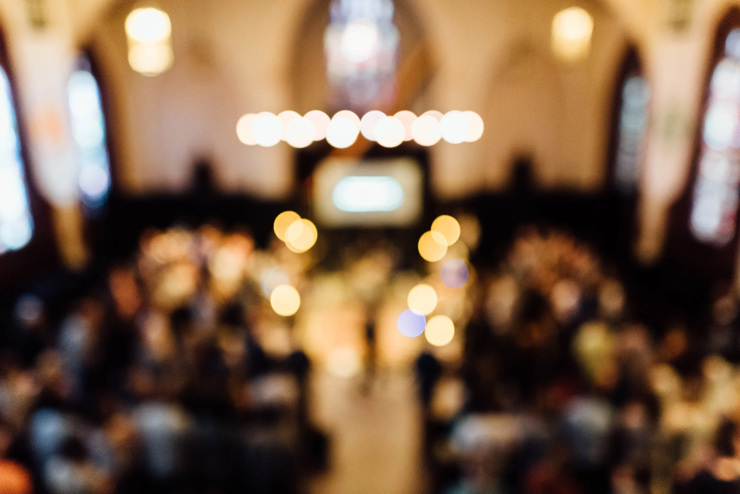 Blurred image of people at a Grace Capital City church worship service in Washington, DC. It shows the church interior with stained glass windows and hanging lighting.