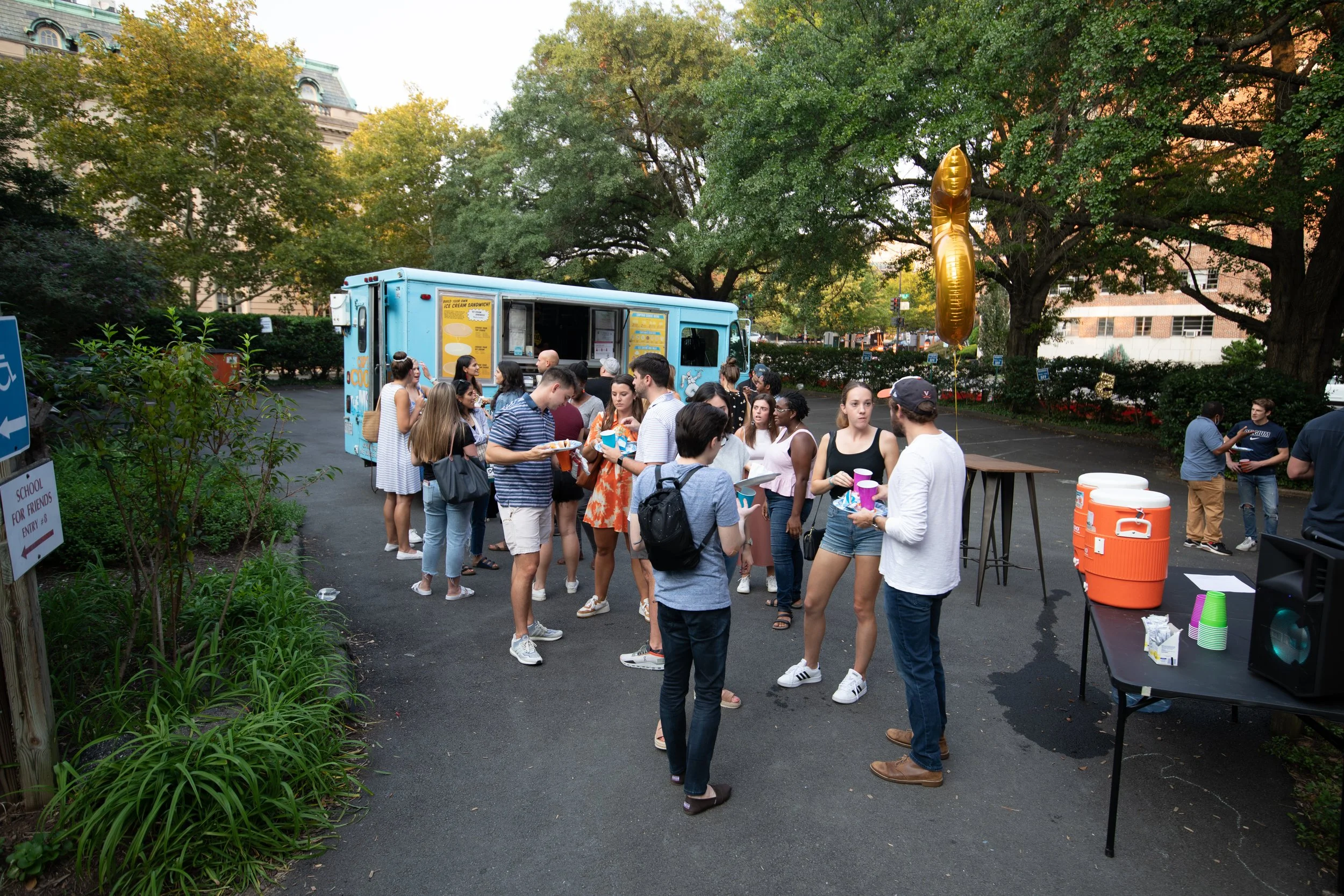 People lined up at a food truck outdoors, with some holding food and drinks, and a large gold balloon in the shape of the number '1' floating above. There are trees and parked cars in the background.